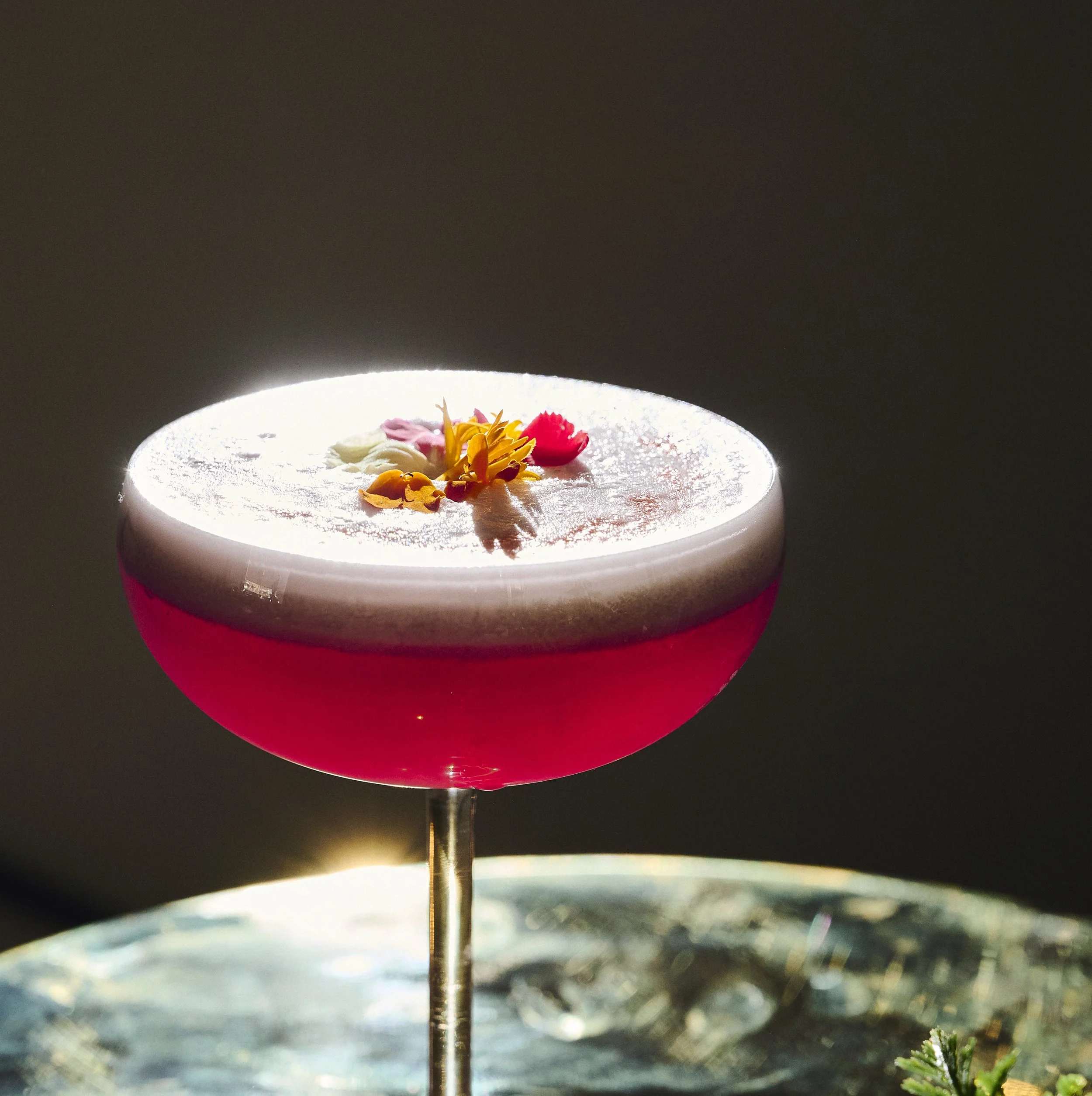 Close-up of a cocktail in a coupe glass with a frothy white top, decorated with edible flowers, set against a dark background.