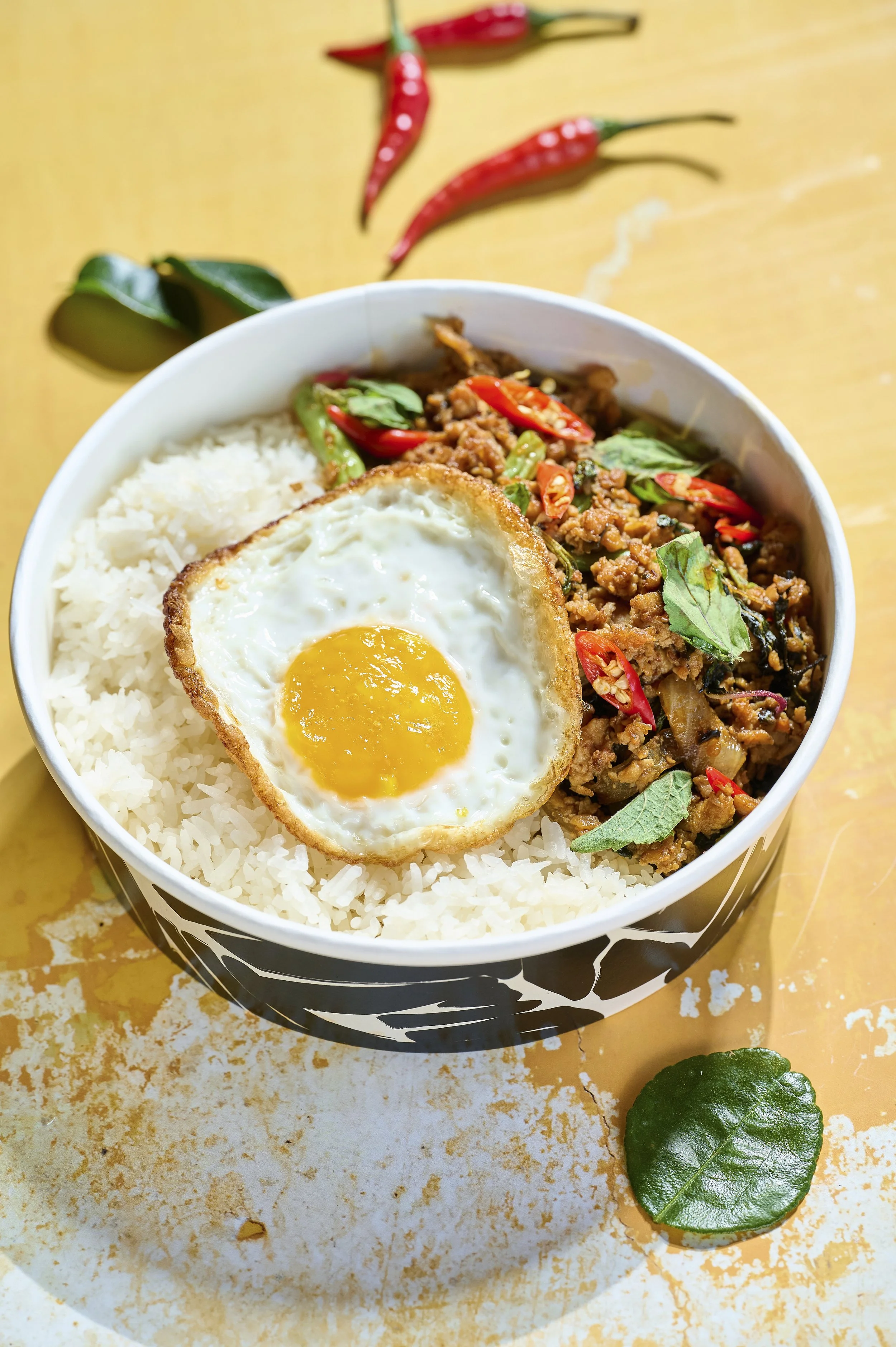 A bowl of white rice topped with a fried egg, spicy stir-fried meat with red chili slices, and fresh green herbs, with red chili peppers and a green leaf on a wooden surface.