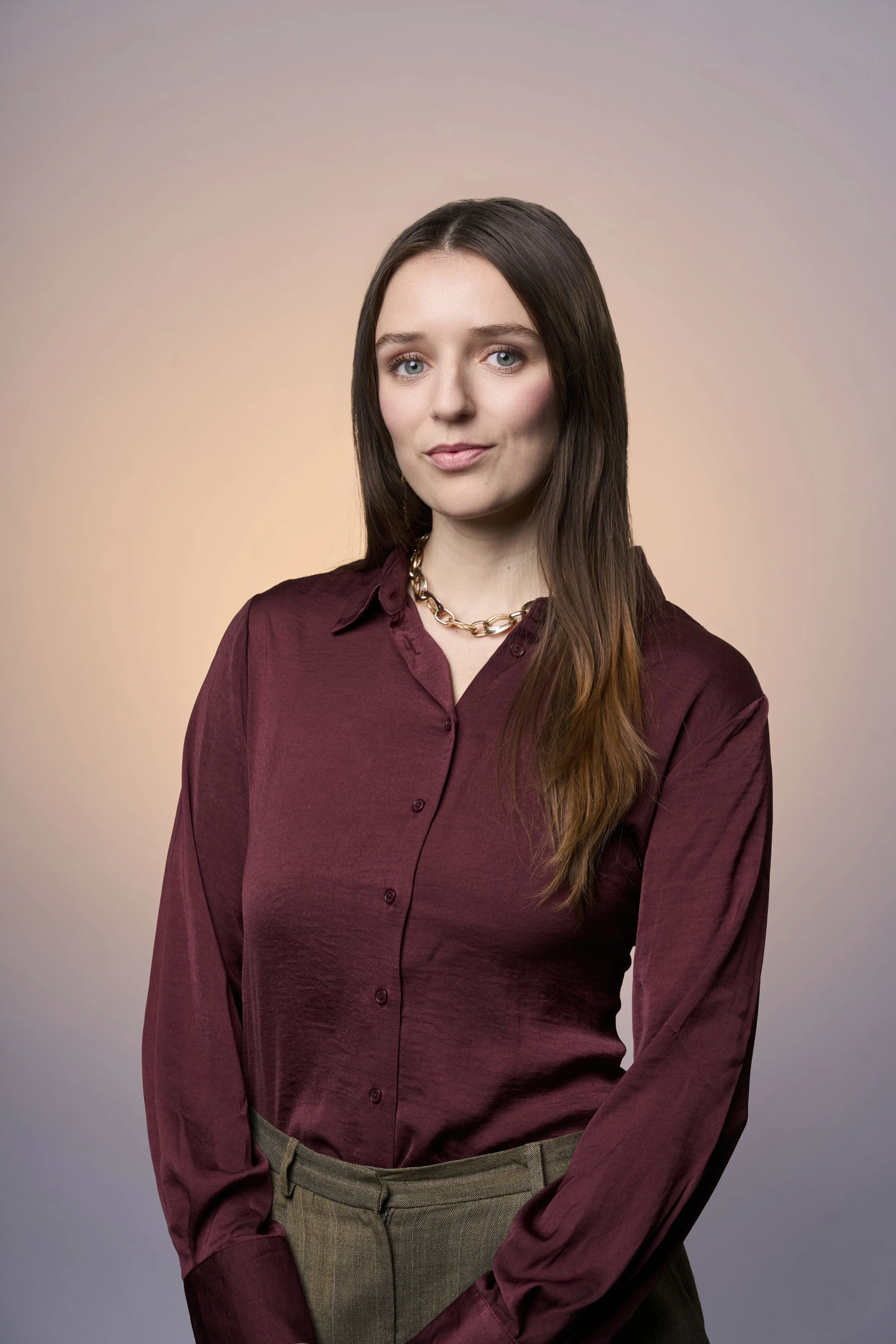 A woman with long brown hair wearing a dark red blouse and a gold chain necklace standing against a neutral gradient background.