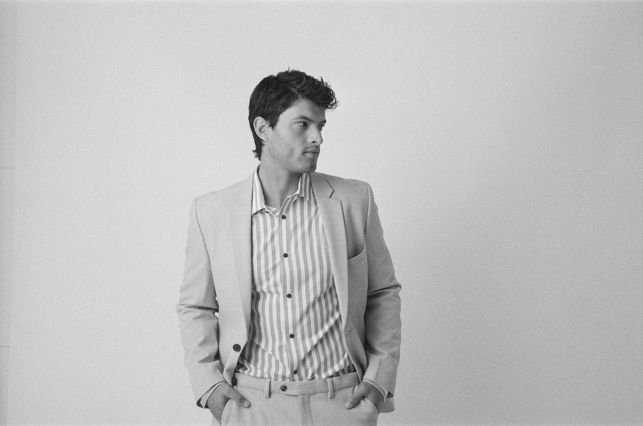 Black and white photo of a young man in a light-colored suit with a striped shirt, standing against a plain wall, looking to the left with hands in pockets.