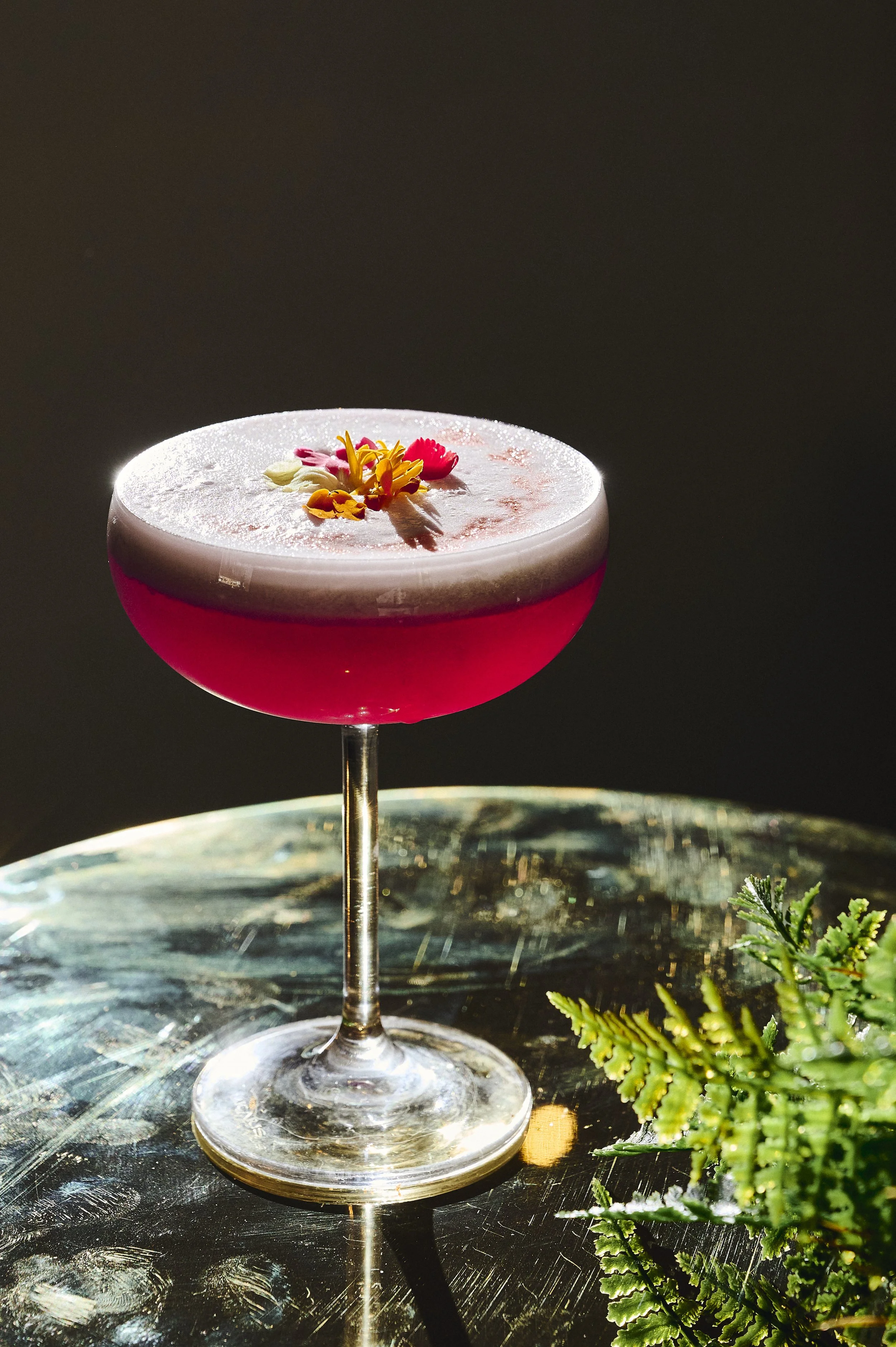 A cocktail in a glass with a wide bowl, filled with a dark pink liquid, topped with a frothy white foam and garnished with colorful edible flowers, placed on a reflective surface with a small green plant beside it.