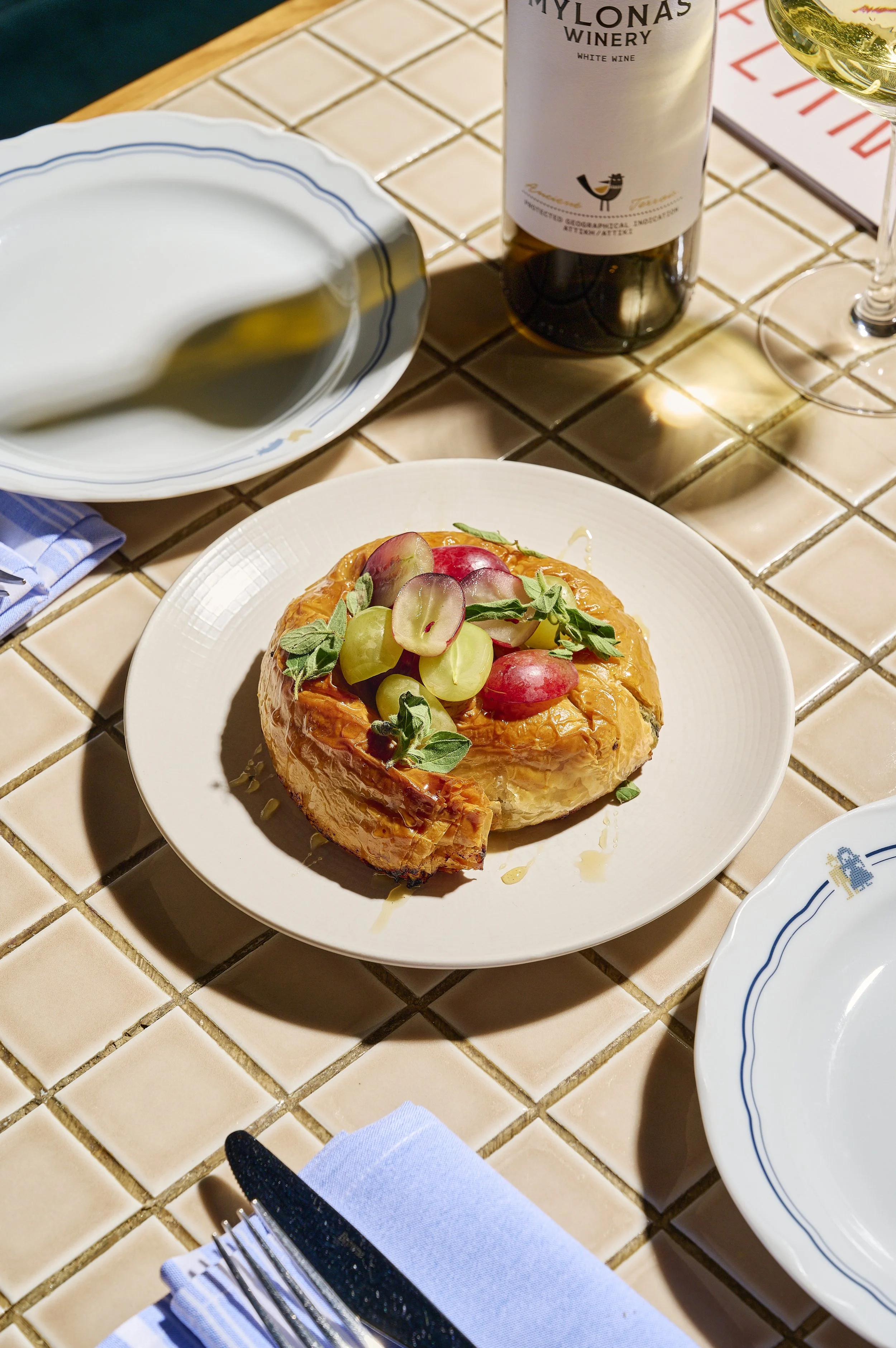 A plate with baked brie cheese topped with green and red grapes and herbs on a tiled table, with a bottle of wine, a glass of white wine, a bowl of olive oil, and a set of silverware and napkin nearby.