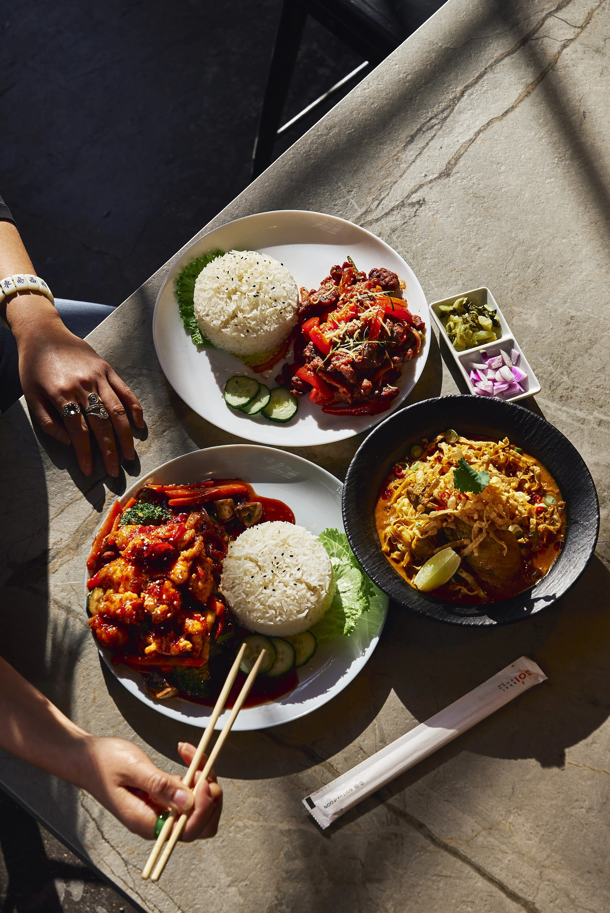 Top-down view of two dishes with rice, meat, and vegetables on white plates, a bowl of noodle soup, small dishes of pickled vegetables, and chopsticks on a stone table.