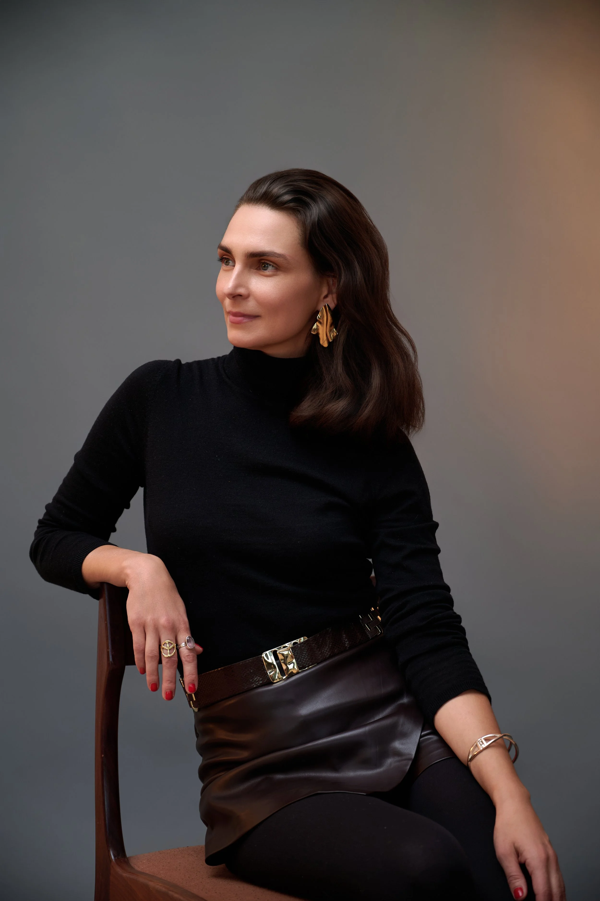 A woman with dark brown hair wearing a black turtleneck, gold earrings, and bracelets, sitting on a brown chair against a gray background.