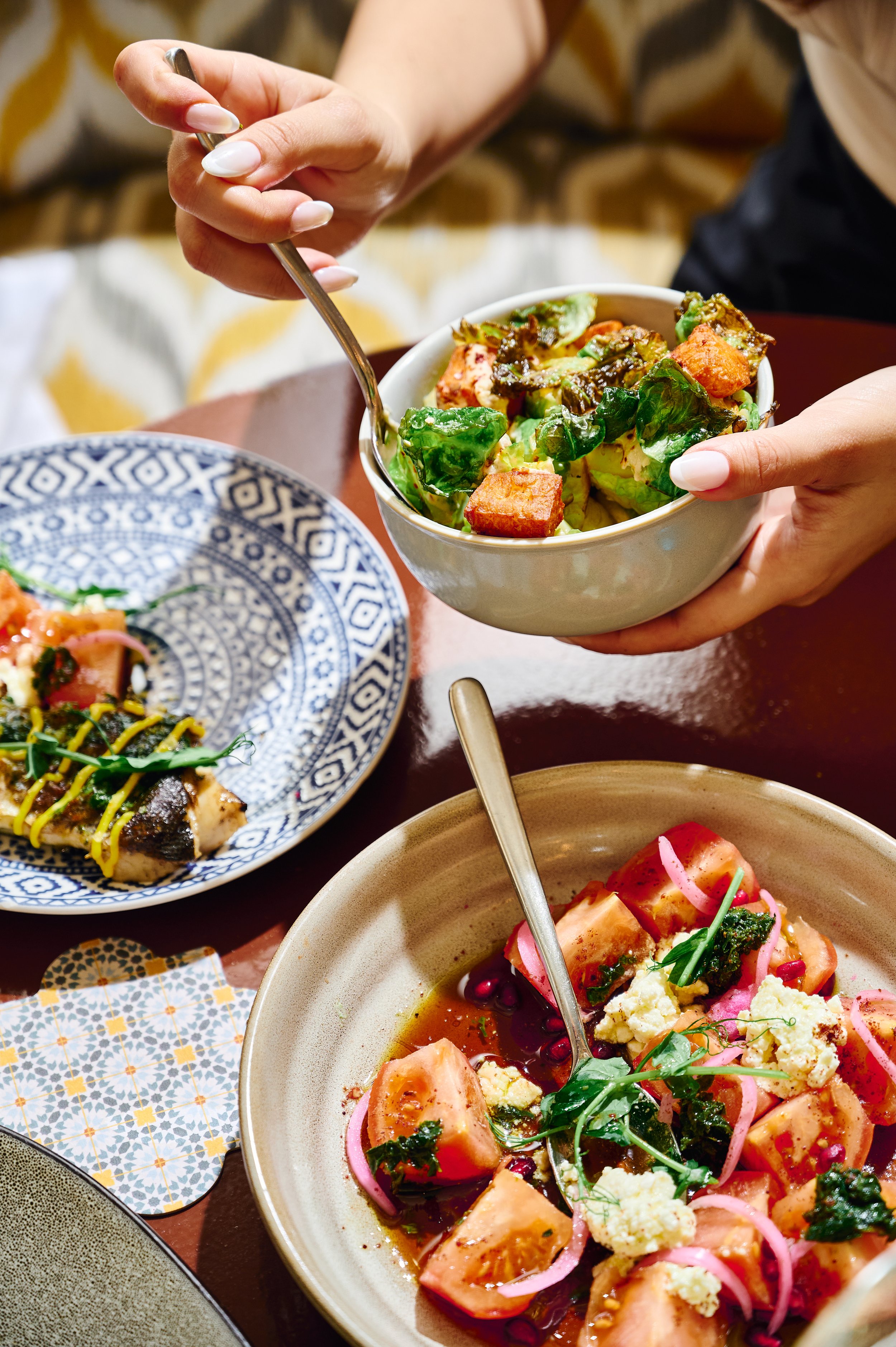 Close-up of a person's hand holding a bowl of salad with greens, tomatoes, and dressing, while another hand holds a fork near the salad. There are two other dishes on the table, one with a fish dish and another with a tomato and onion salad garnished