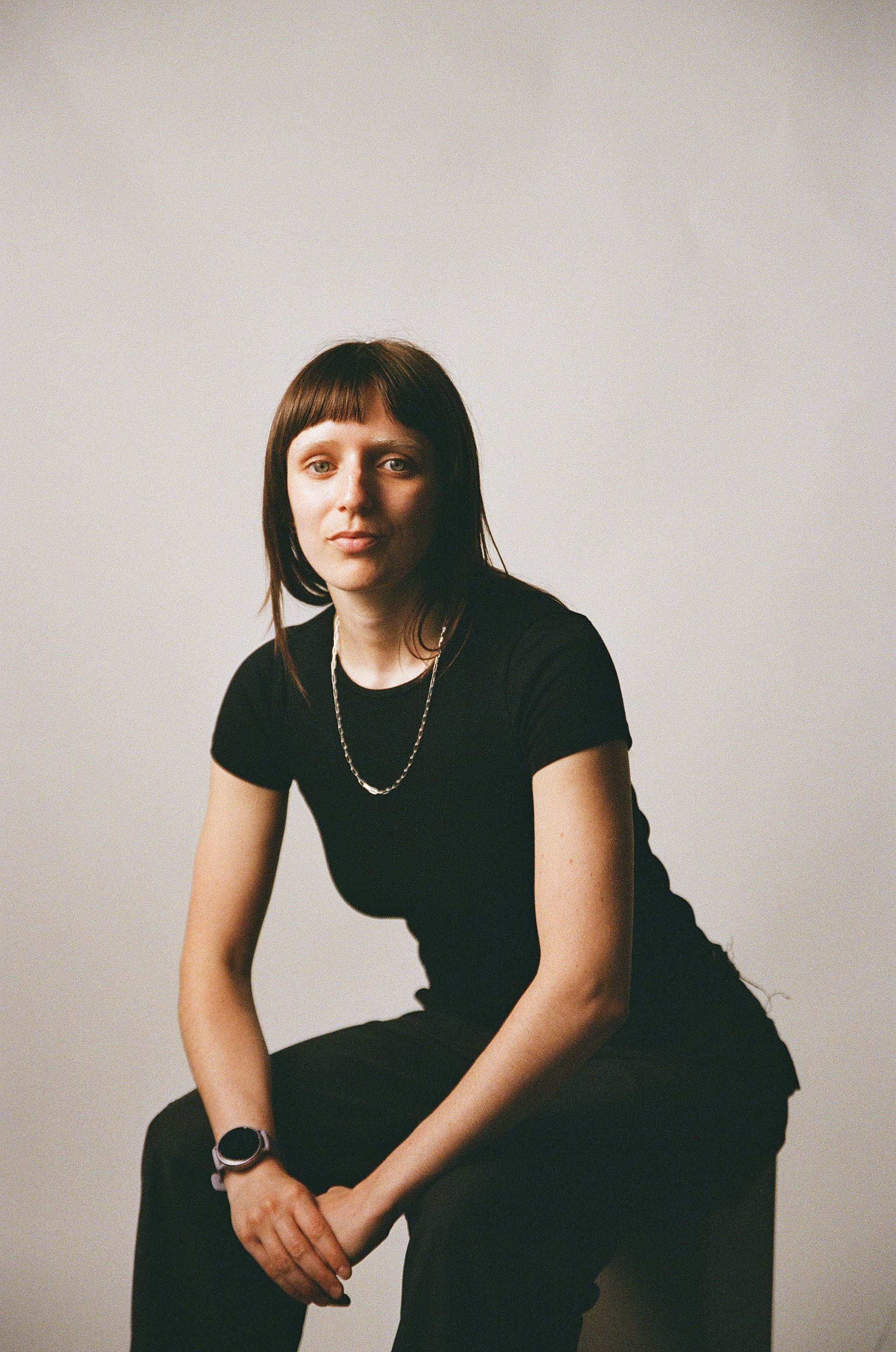 Portrait of a woman with shoulder-length brown hair and bangs, wearing a black t-shirt, a silver necklace, and a smartwatch, seated against a plain background.