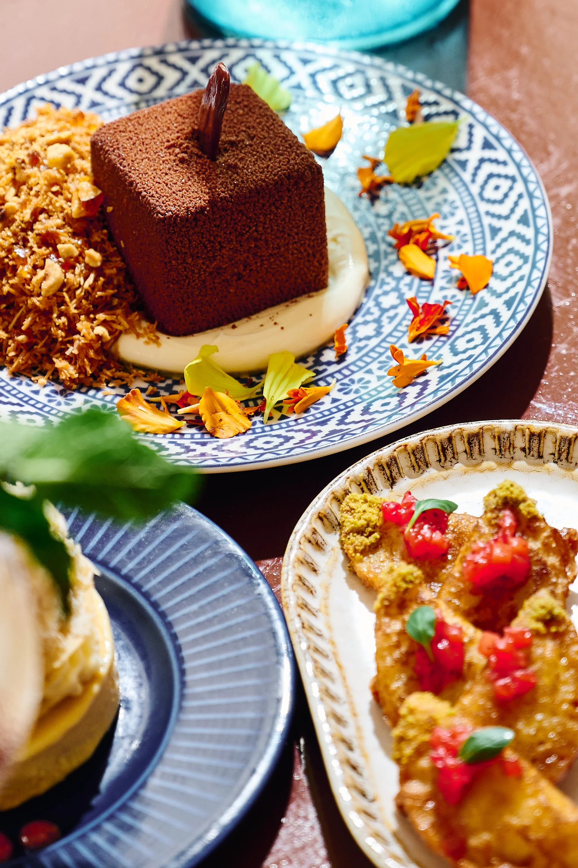 Assorted Indian dishes on decorative plates, including a chocolate dessert, a fried item garnished with red sauce and herbs, and a rice dish with nuts, served on a wooden table.