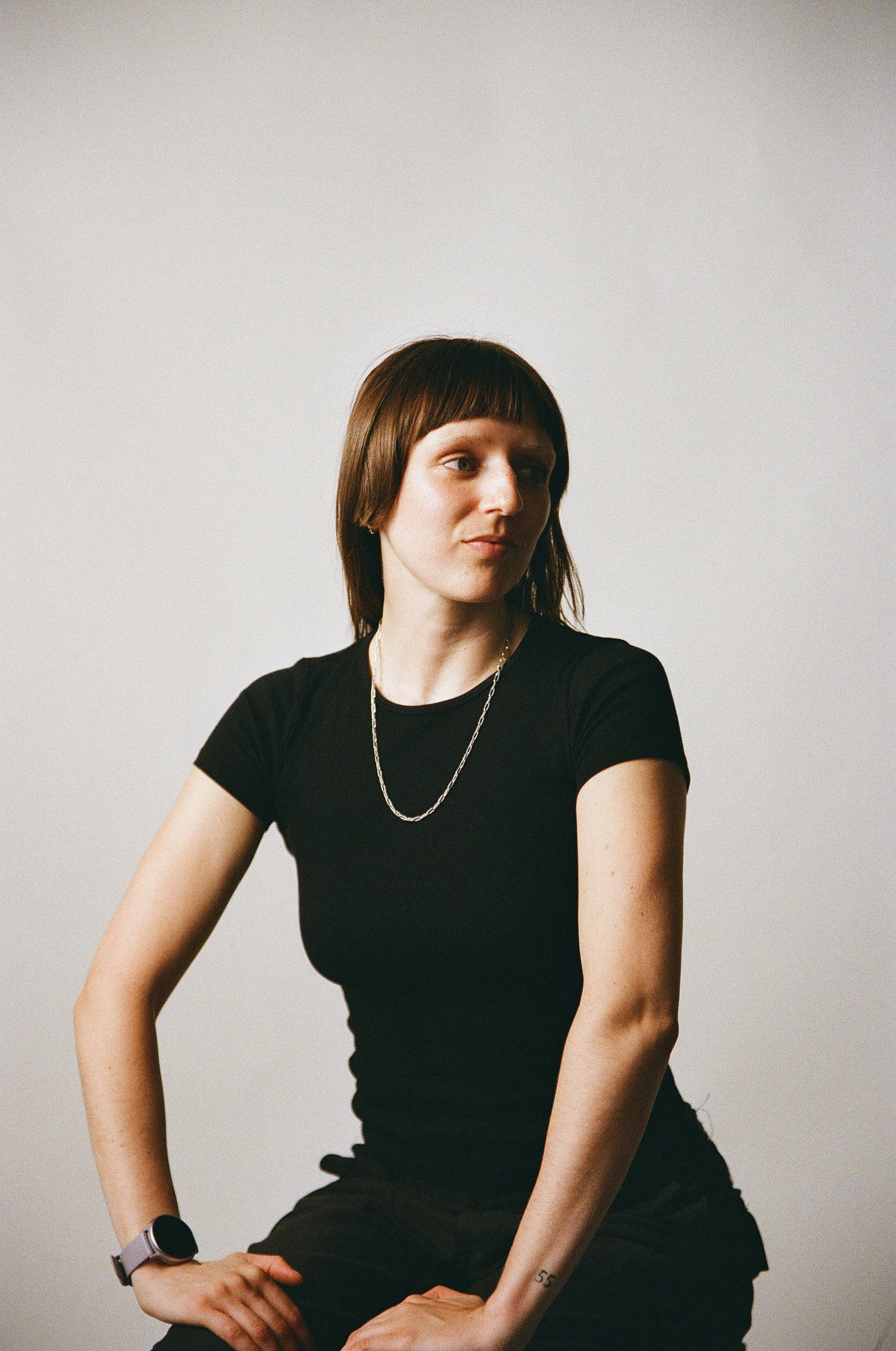 A woman with shoulder-length brown hair and bangs, wearing a black t-shirt, a silver chain necklace, and a smartwatch, sitting against a plain light-colored background.