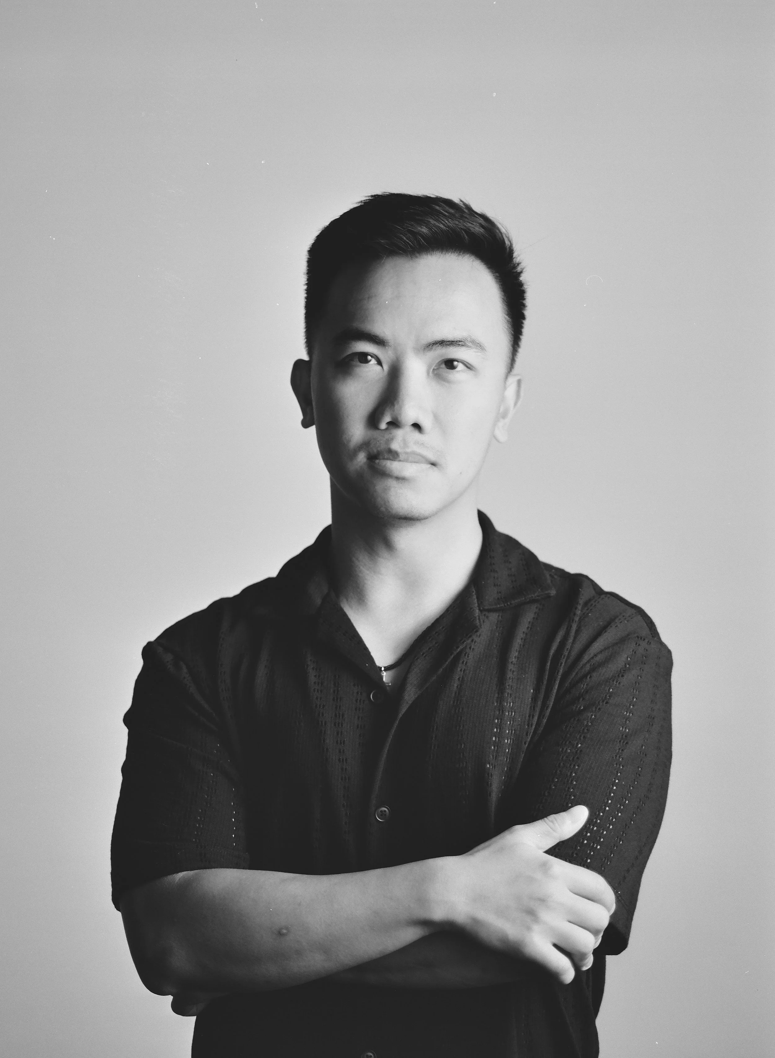 Black and white portrait of a young man with short hair, wearing a dark shirt with rolled-up sleeves, standing against a plain background with arms crossed.