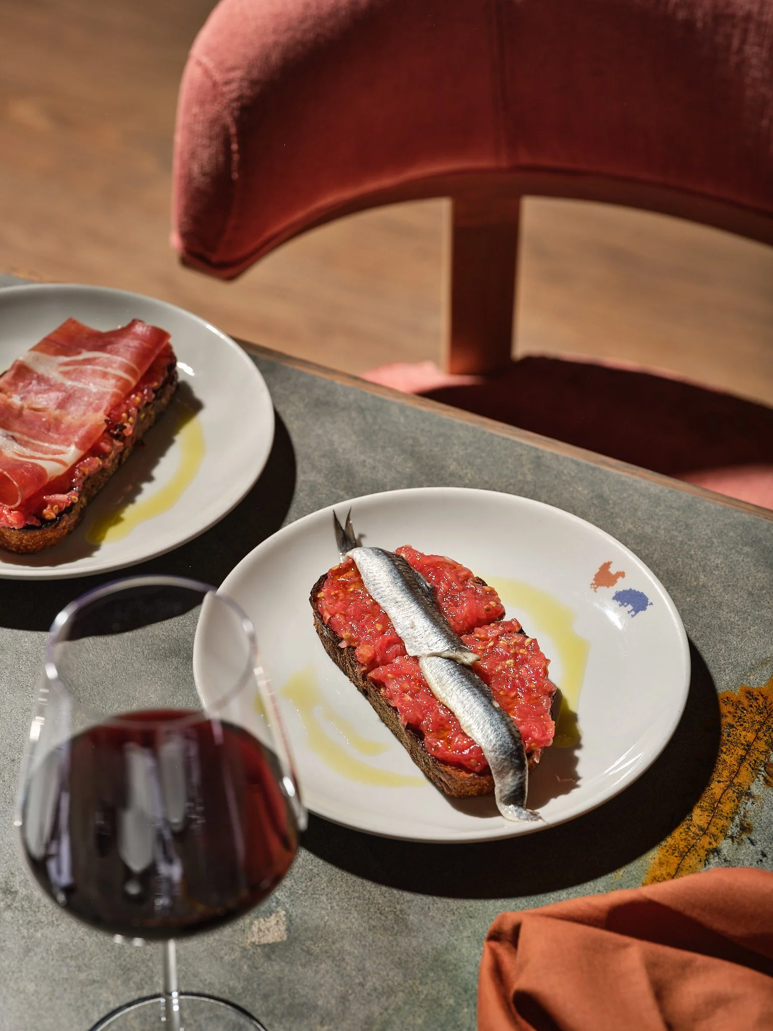 Two plates of bruschetta with tomato topping and anchovies, a glass of red wine, and a pink upholstered chair in the background.