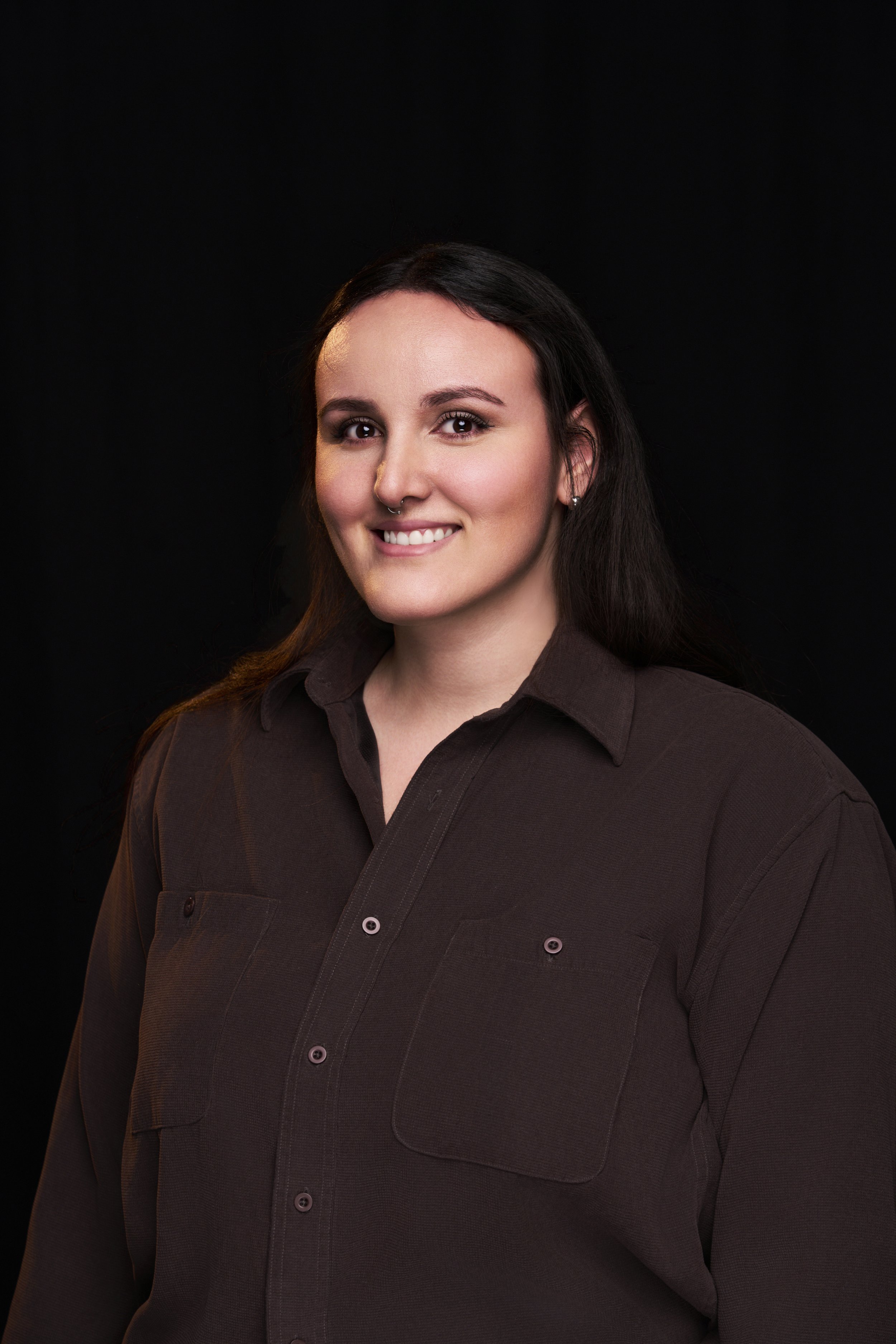 A smiling woman with dark hair and a septum piercing, wearing a brown button-up shirt, against a black background.