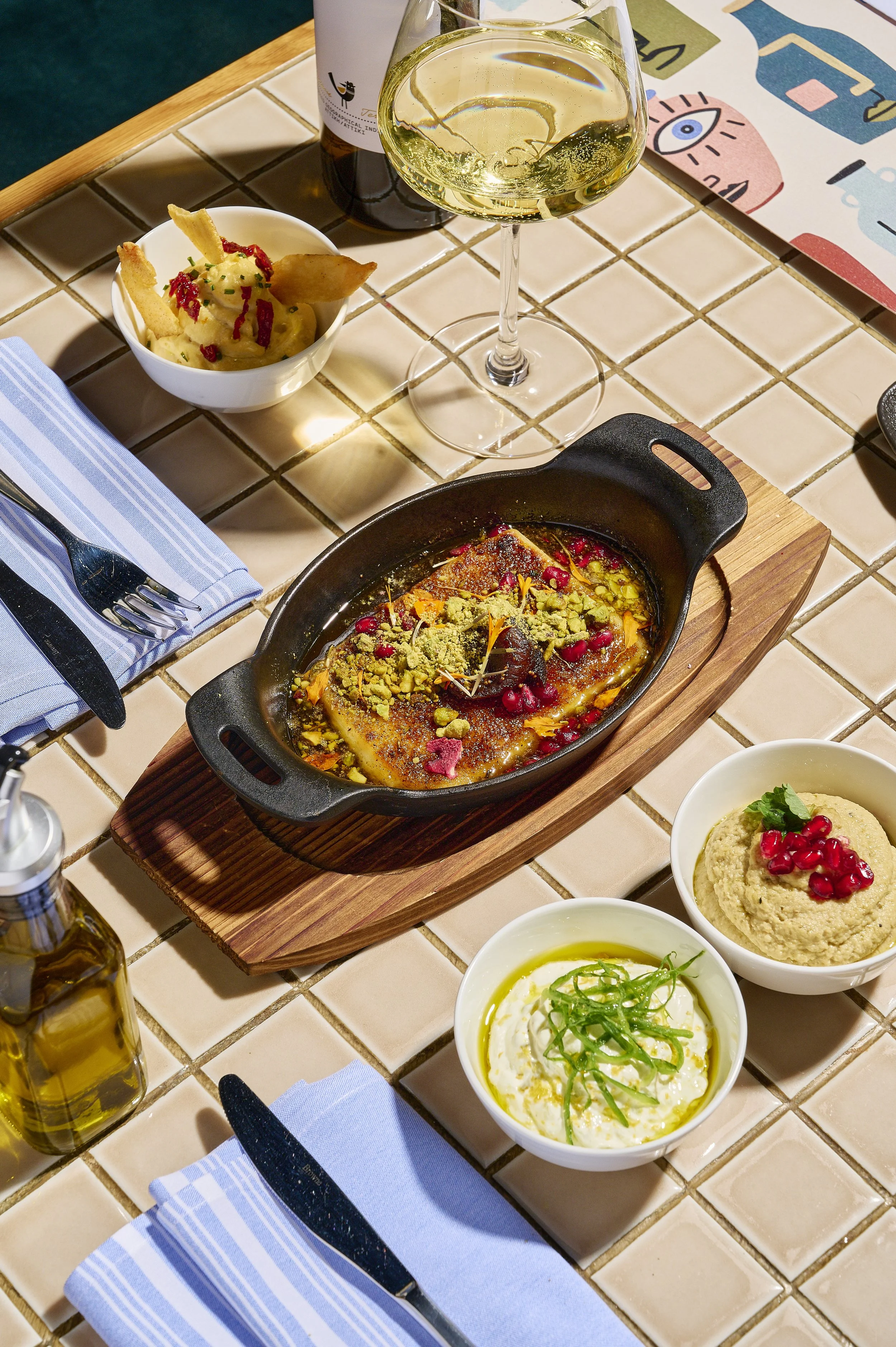 A meal on a tiled table includes a small pan of baked fish topped with herbs and pomegranate seeds, a bowl of mashed potatoes with pomegranate, a bowl of creamy dip garnished with green herbs and oil, and a bowl of hummus with pomegranate on top. The