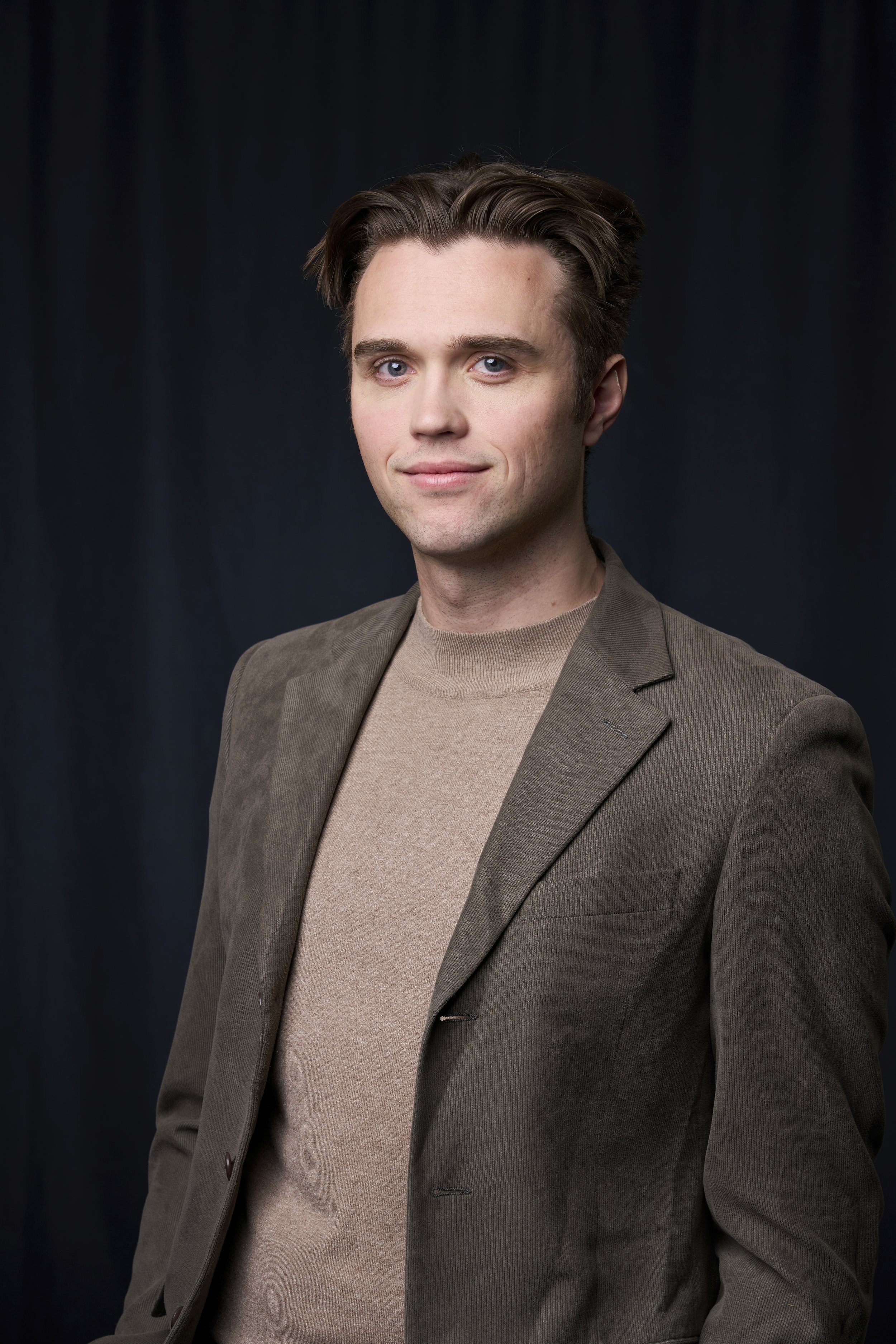 Portrait of a young man with brown hair and blue eyes wearing a beige turtleneck and a brown blazer against a dark background.