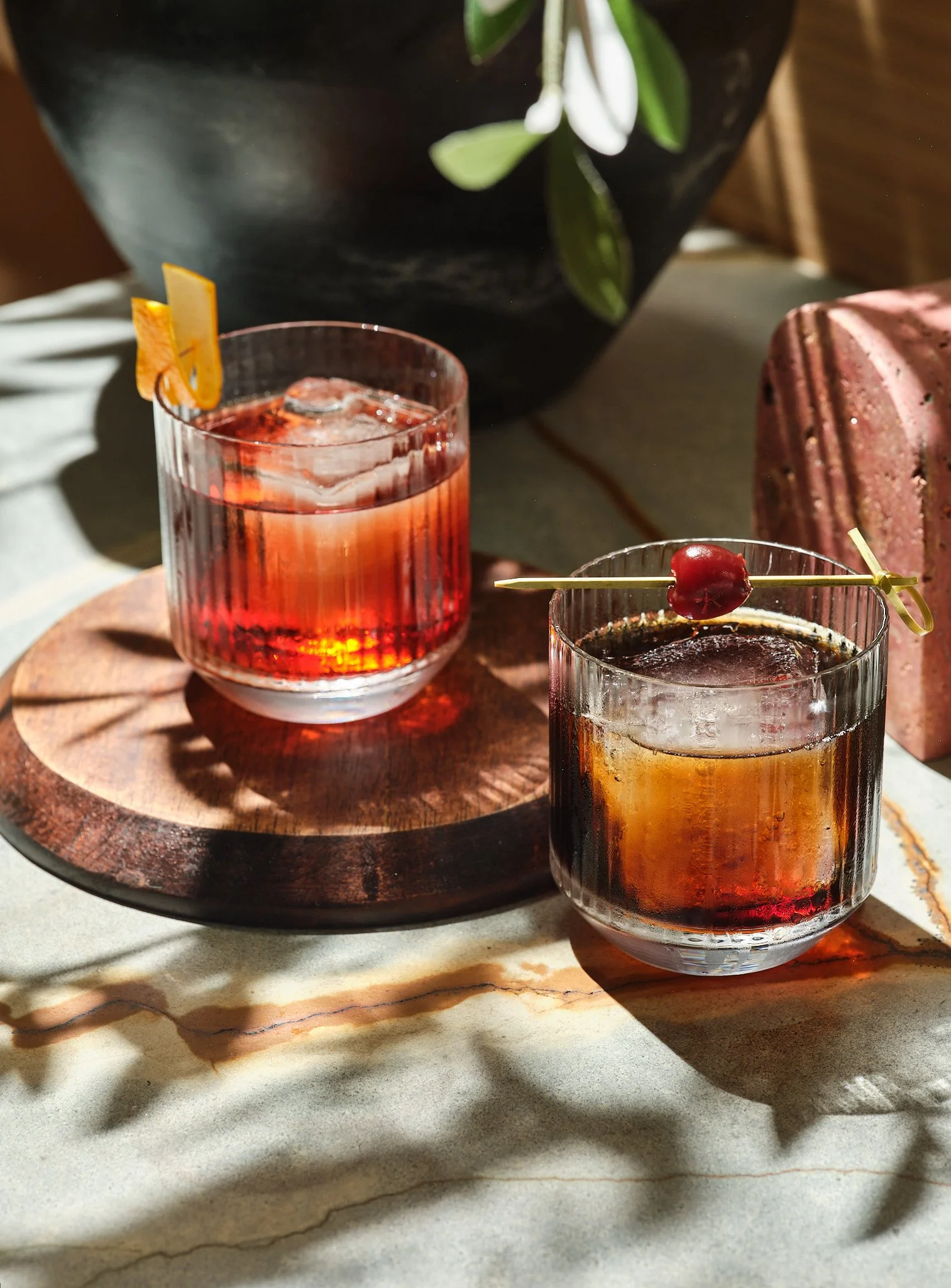 Two cocktails with ice, one garnished with a cherry on a skewer and the other with a citrus peel, placed on a wooden serving board near a marble surface.