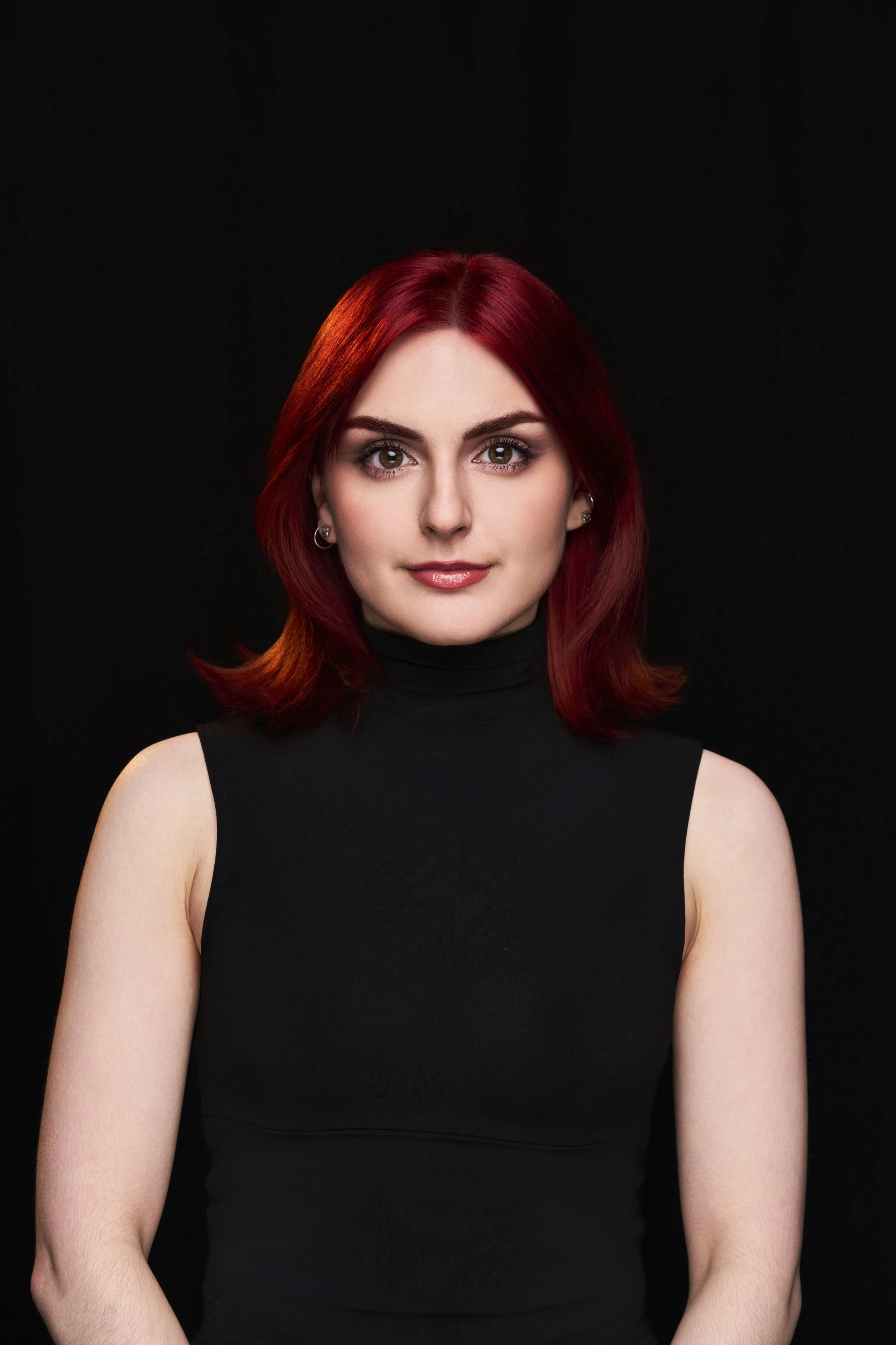 A woman with red hair and a black sleeveless turtleneck against a black background.