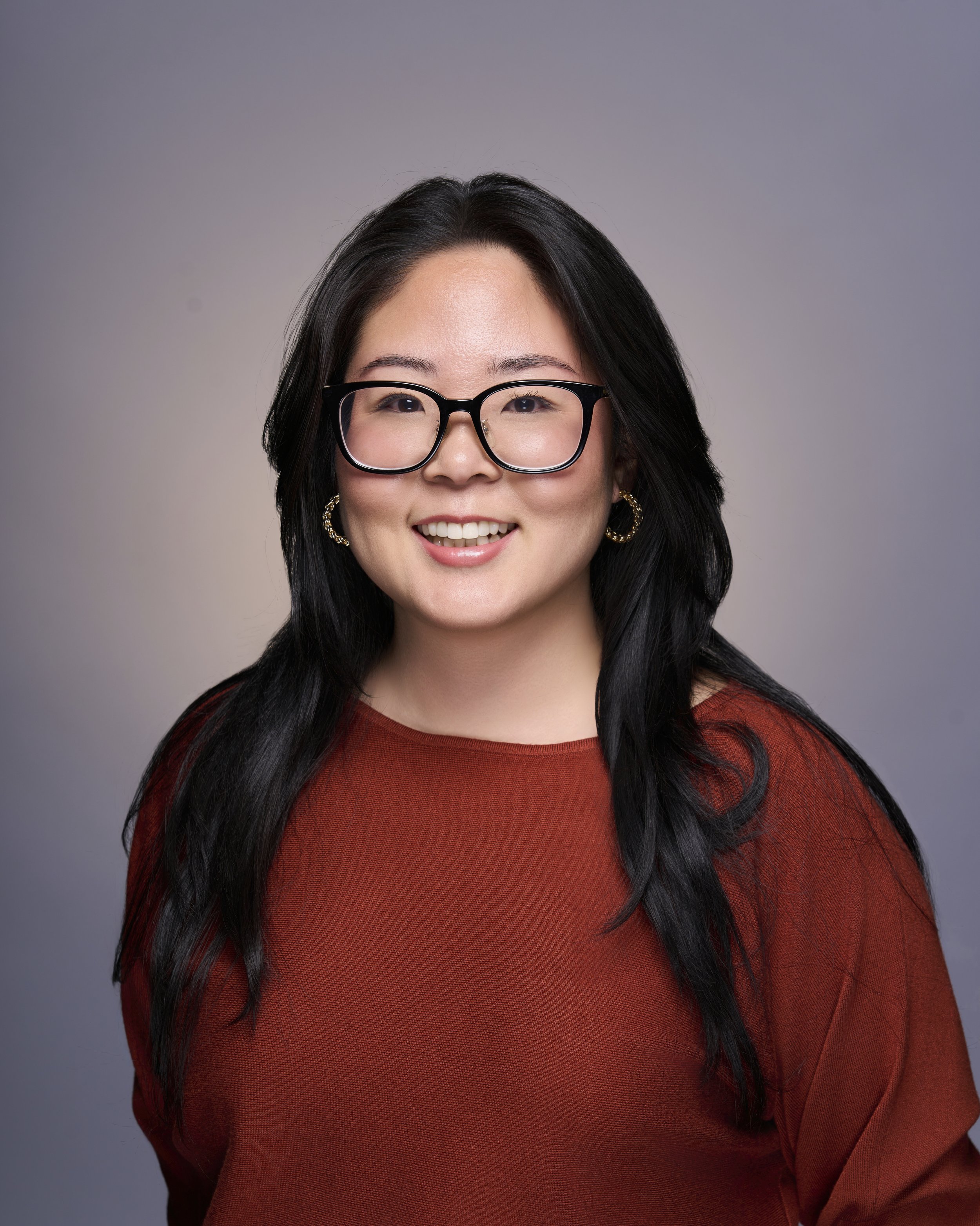 A woman with long black hair, wearing glasses, gold hoop earrings, and a rust-colored top, smiling against a neutral background.