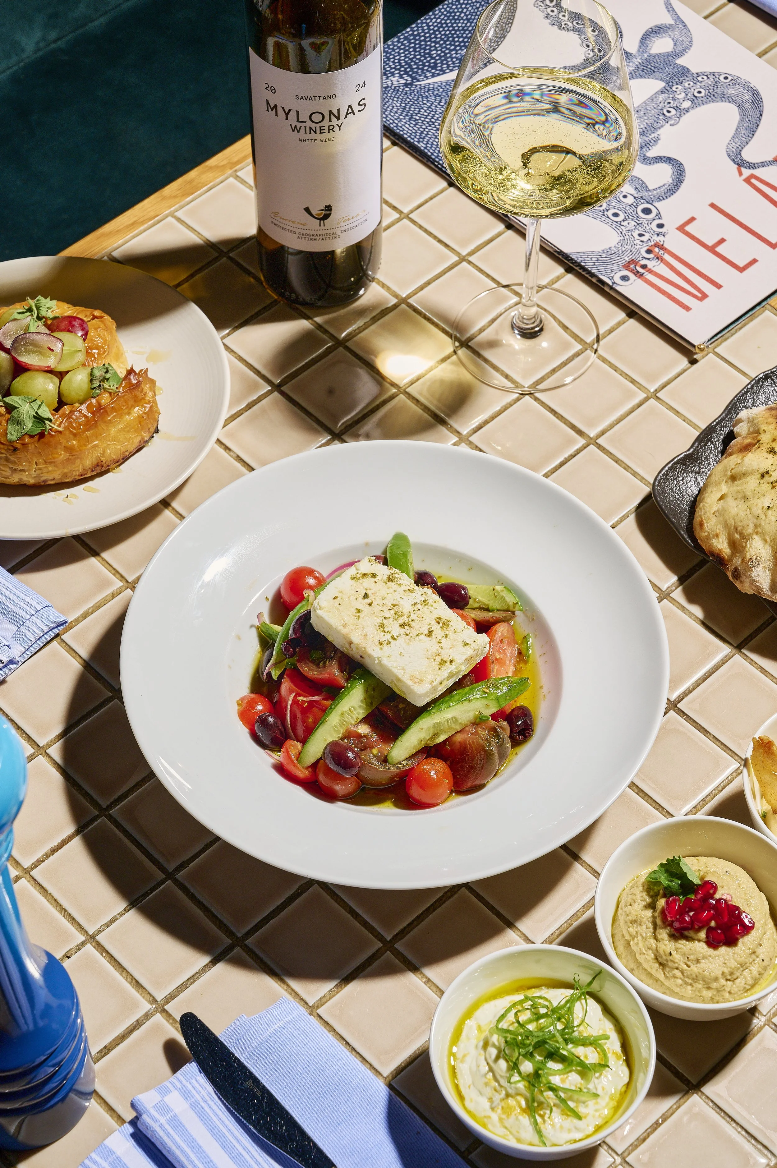 A dining table with a variety of dishes including a plate with salad topped with a block of cheese, bowls of hummus and a dip with pomegranate seeds, a bottle of white wine, a glass of white wine, and a basket of bread. The table has a tiled surface.