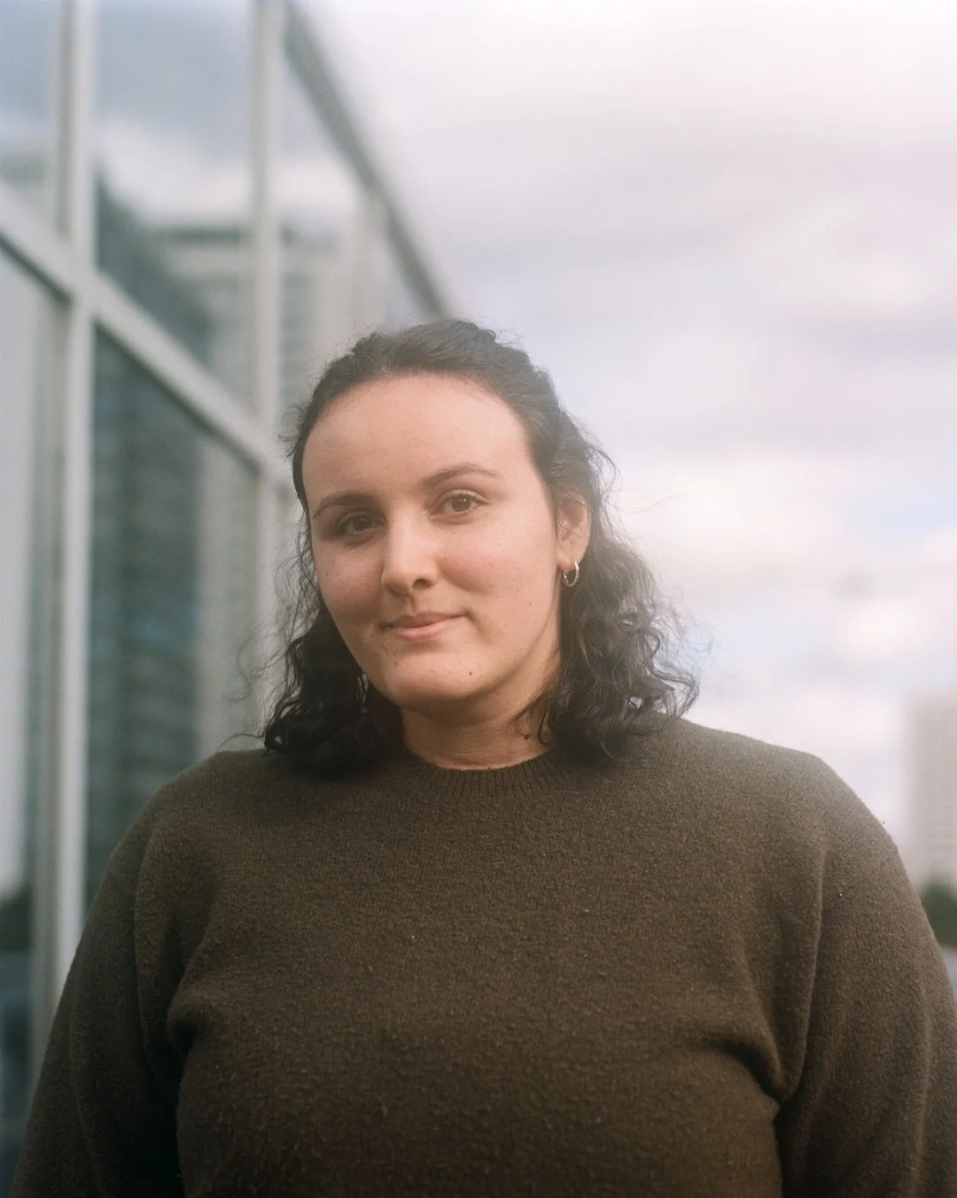A woman with dark curly hair wearing a brown sweater standing outdoors in front of a modern building with glass windows, under a cloudy sky.