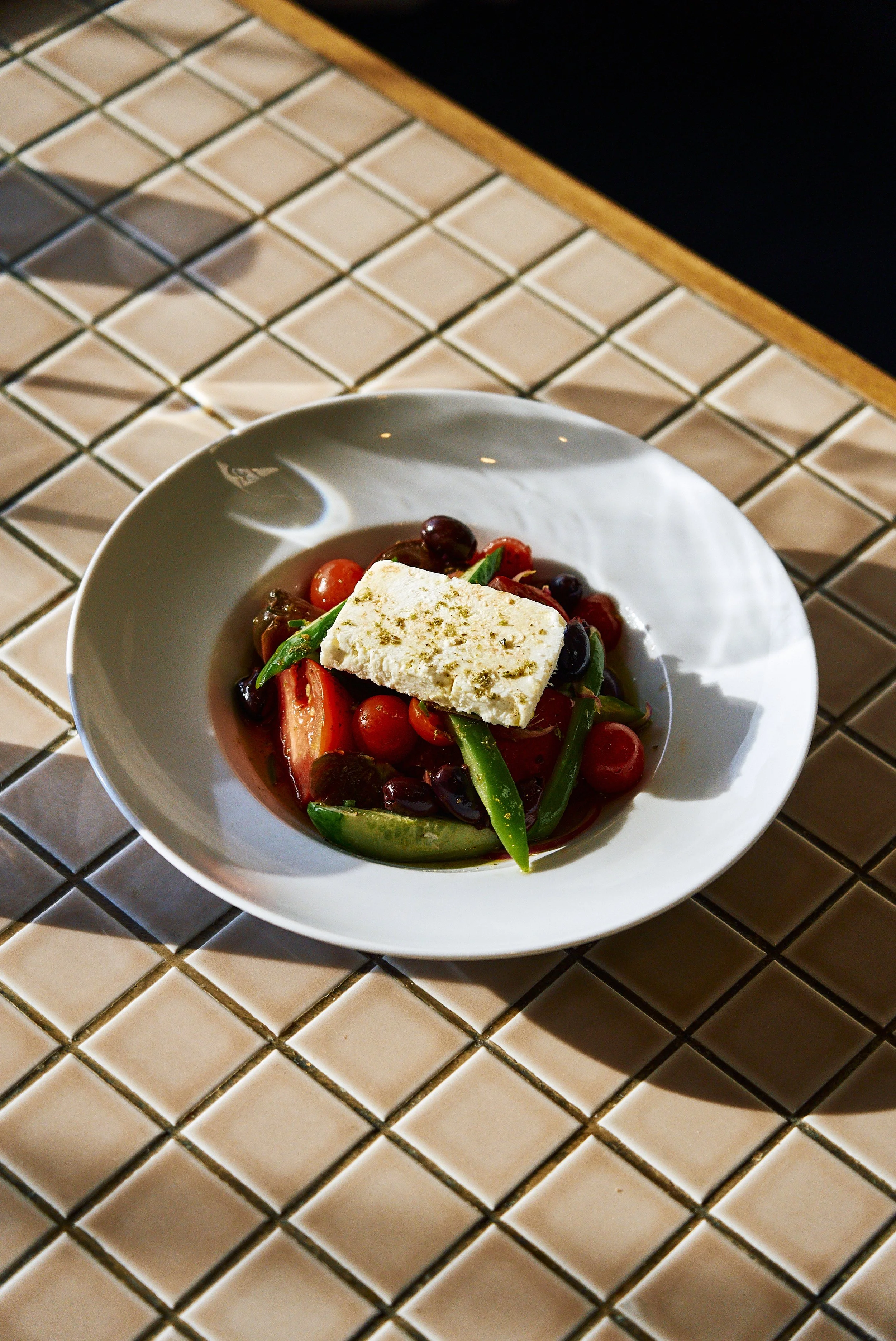 A white bowl of salad with cherry tomatoes, green beans, kalamata olives, topped with a rectangular piece of cheese, on a tiled table.