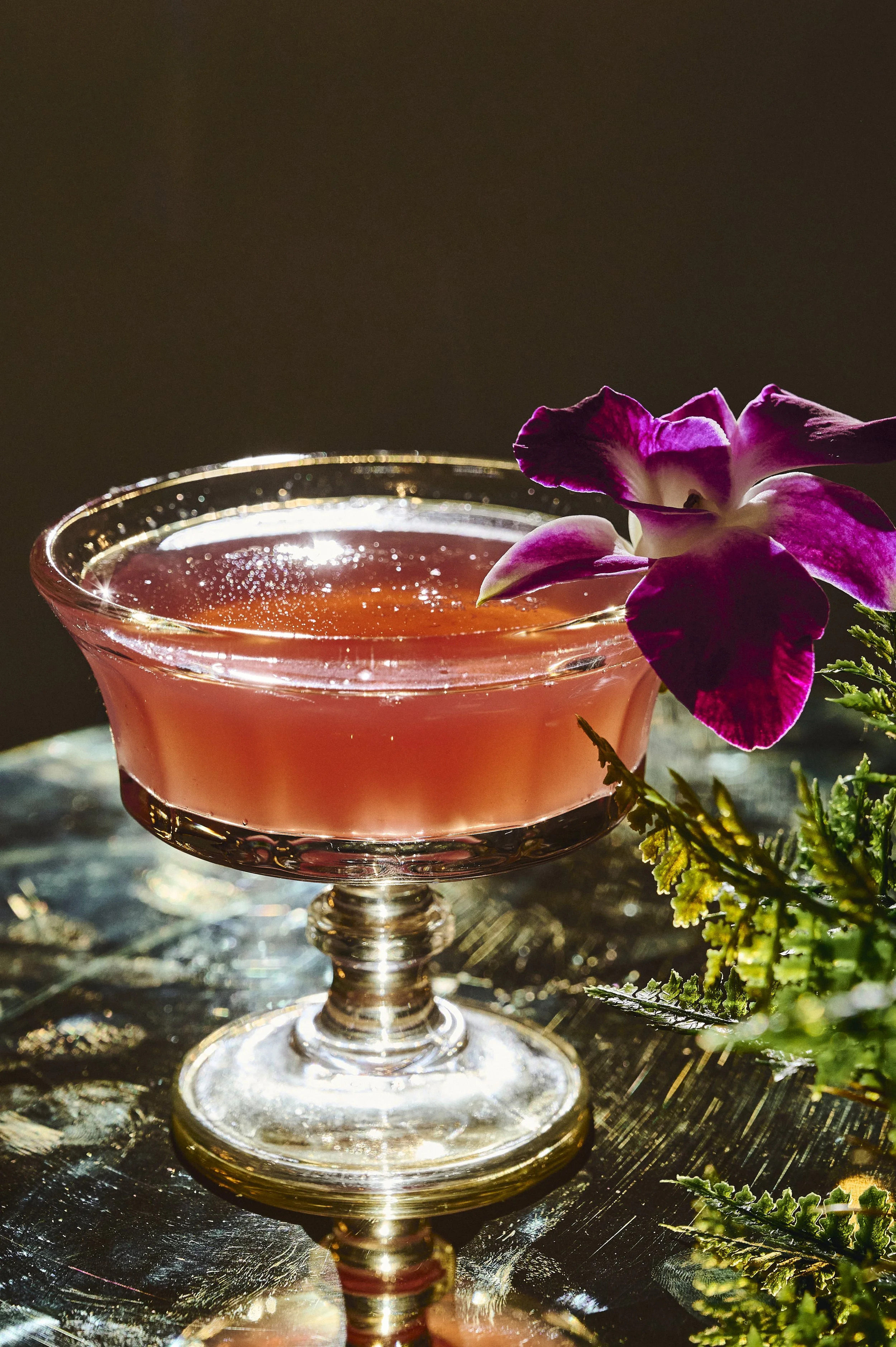 A pink cocktail in a glass with a purple orchid flower garnishing the rim, placed on a reflective surface with greenery nearby.