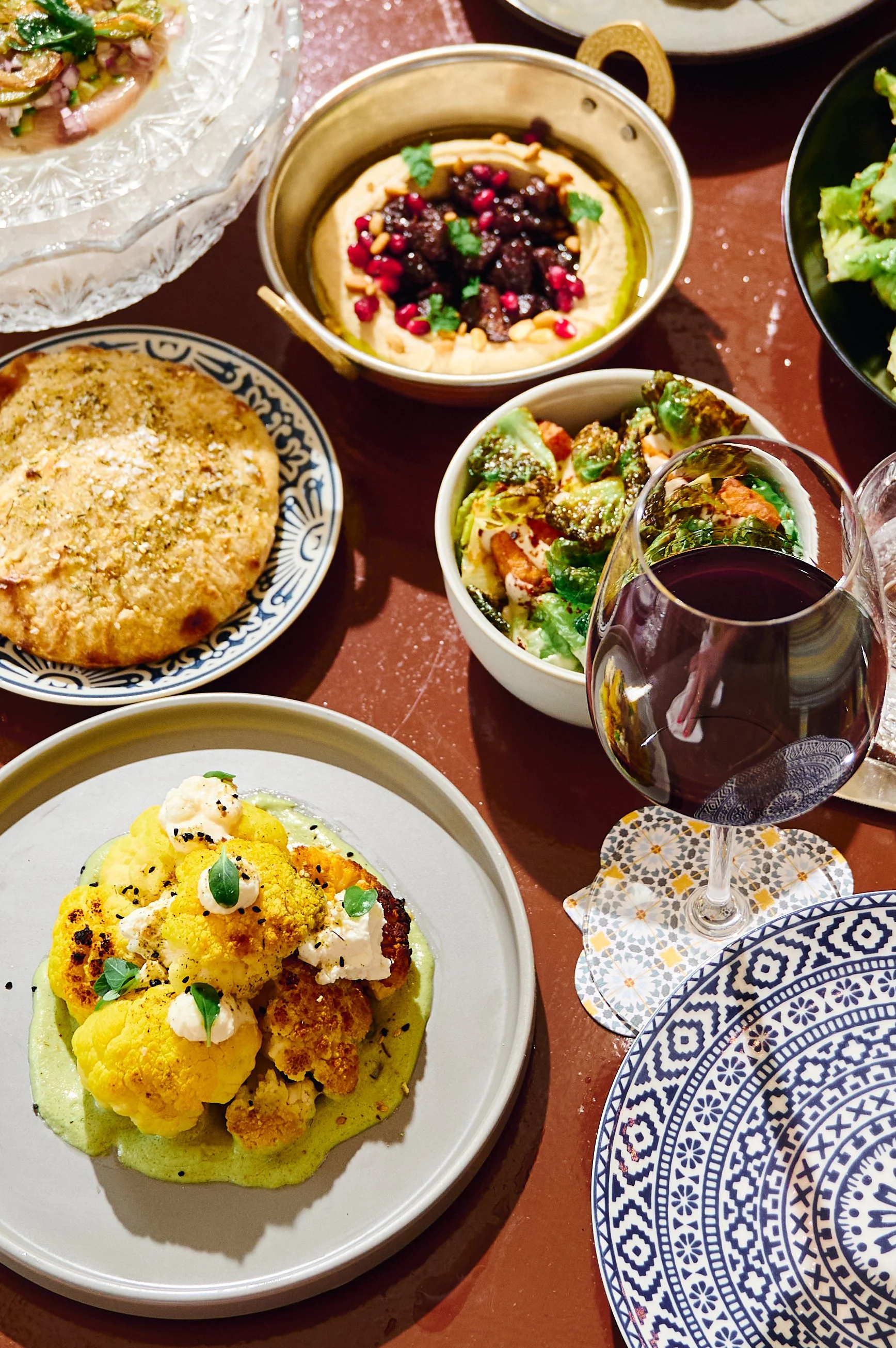 Assorted dishes on a table including cauliflower, a bowl of hummus with toppings, a salad, bread, and a glass of red wine.