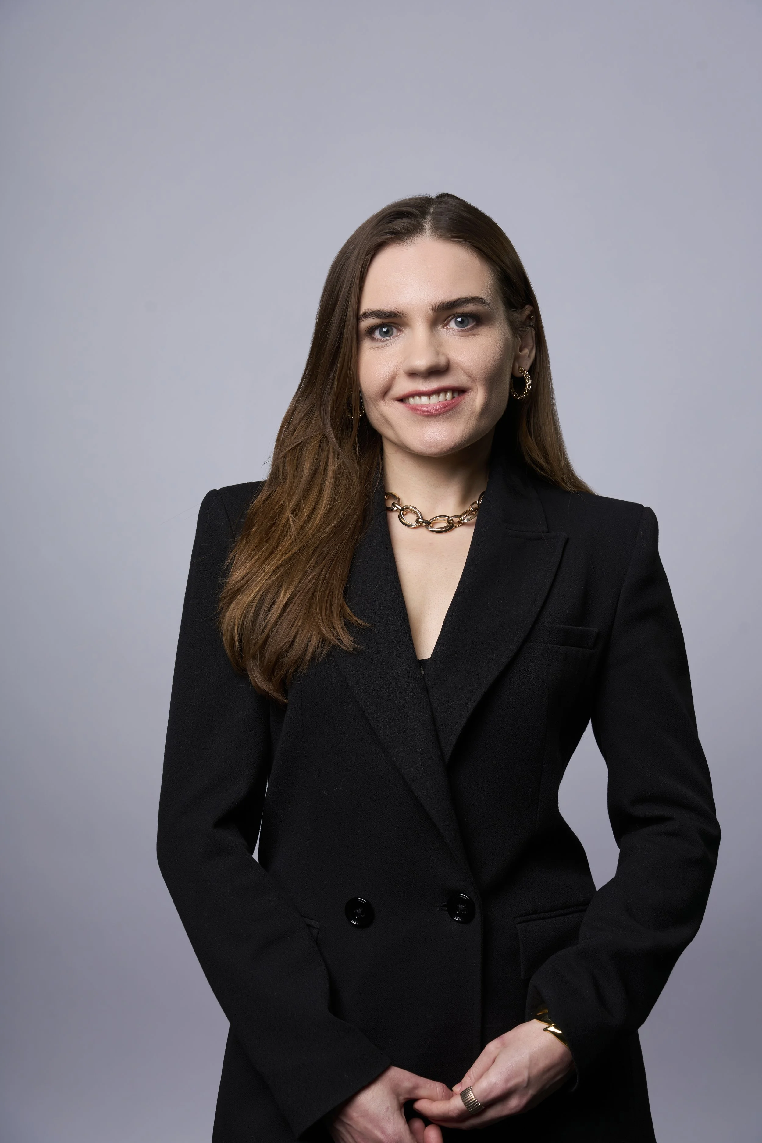 A young woman with long brown hair wearing a black blazer, gold jewelry, and smiling against a plain gray background.