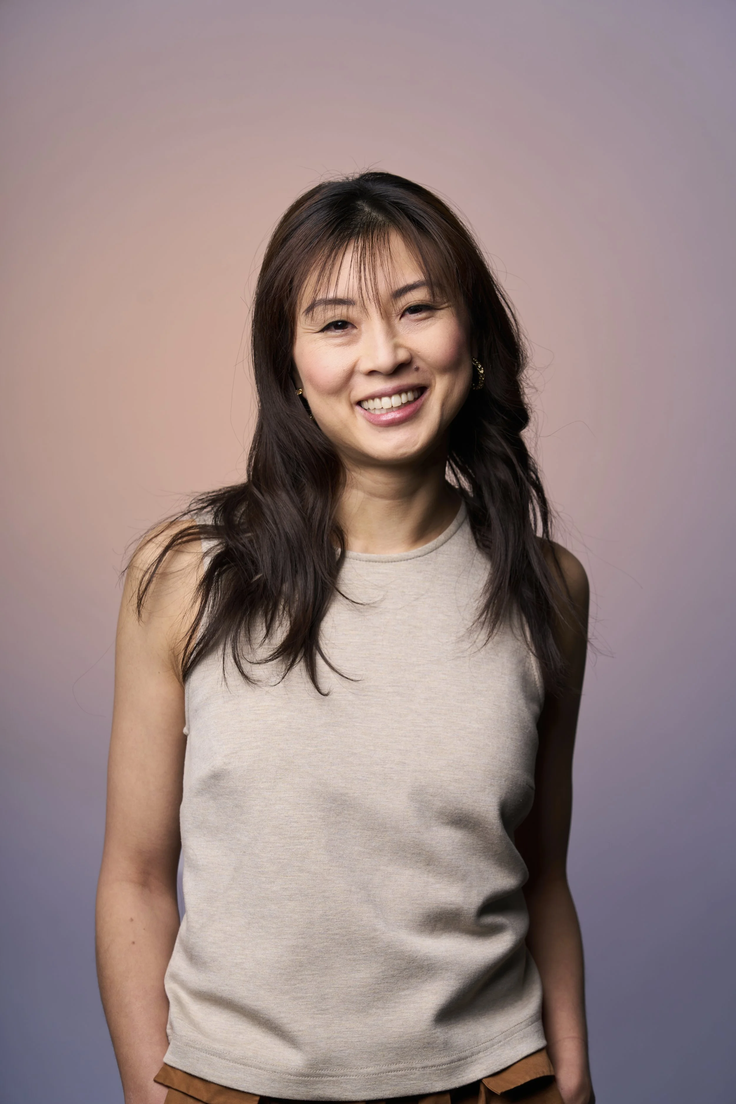 A woman with long dark hair, smiling, wearing a sleeveless beige top, standing against a pastel gradient background.