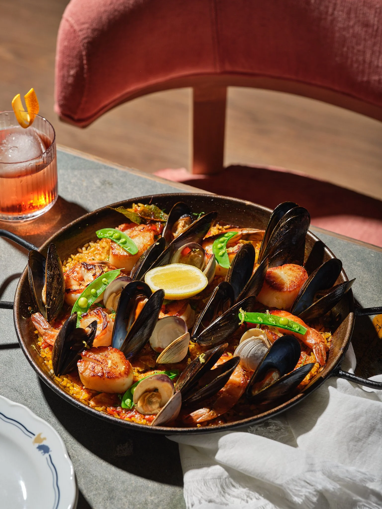 Seafood paella with mussels, scallops, shrimp, and a lemon wedge served in a black skillet on a table with a glass of pink drink and a white plate nearby.