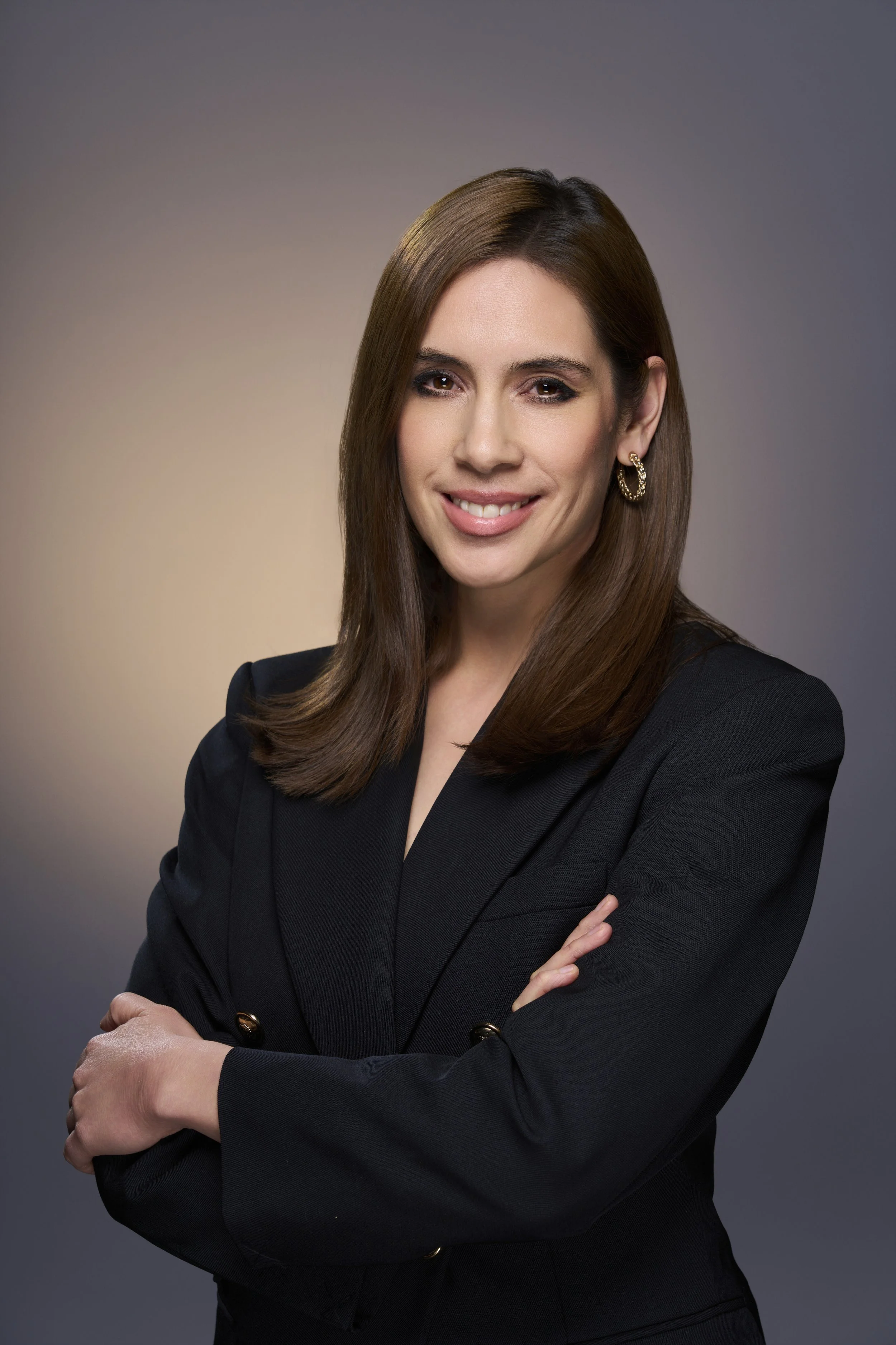 Professional woman with brown hair and gold hoop earrings, wearing a black blazer, smiling with arms crossed against a gradient background.
