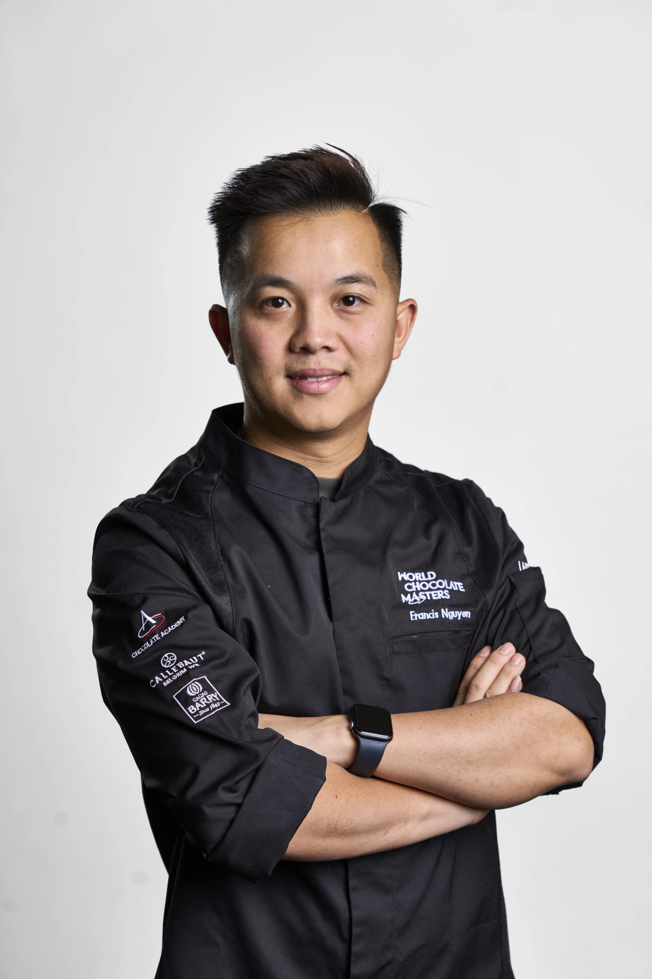 A young man wearing a black chef's coat with embroidered text, standing with arms crossed and smiling in front of a plain white background.