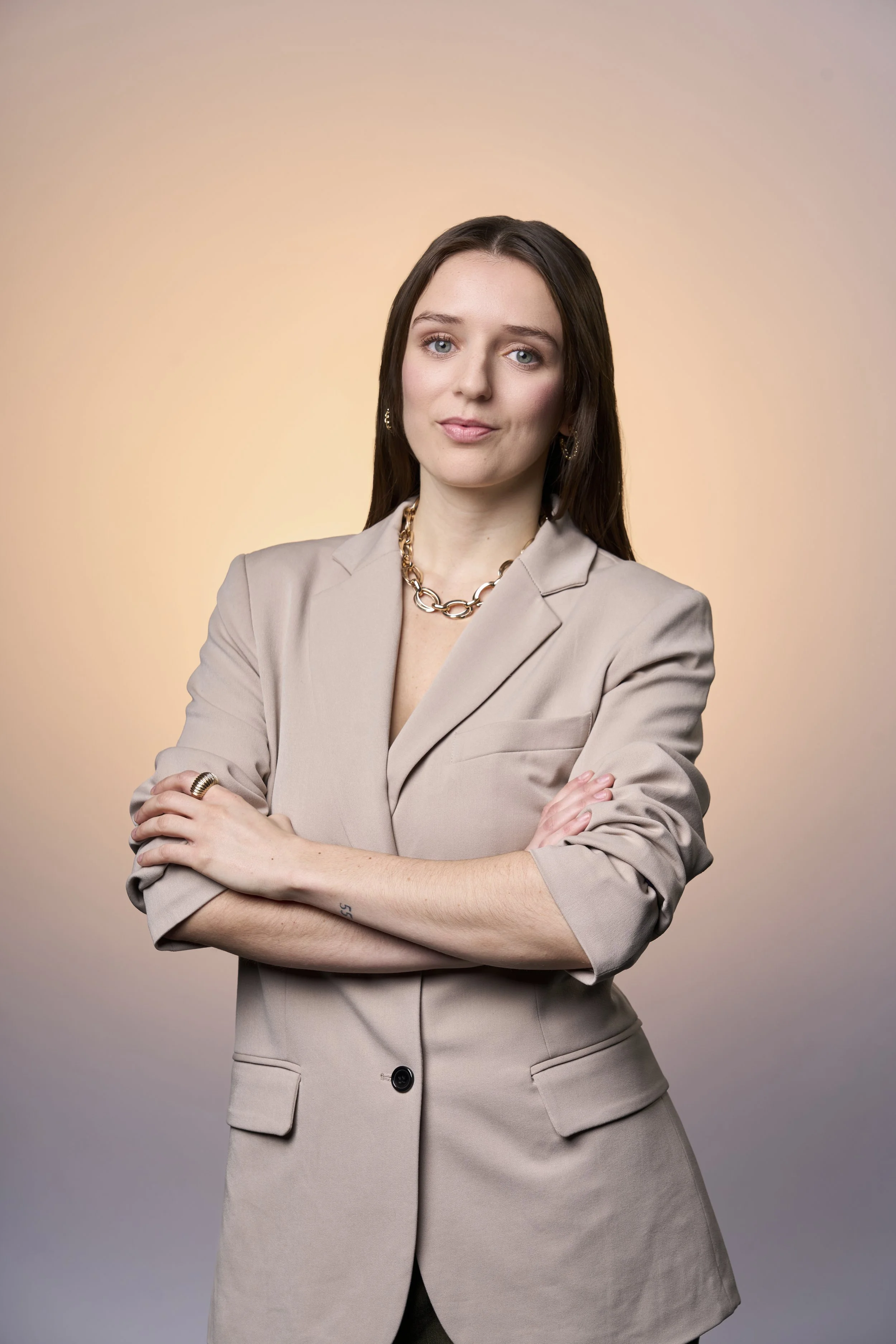 A woman with long brown hair in a beige blazer, gold jewelry, and crossed arms, posing against a gradient peach and gray background.