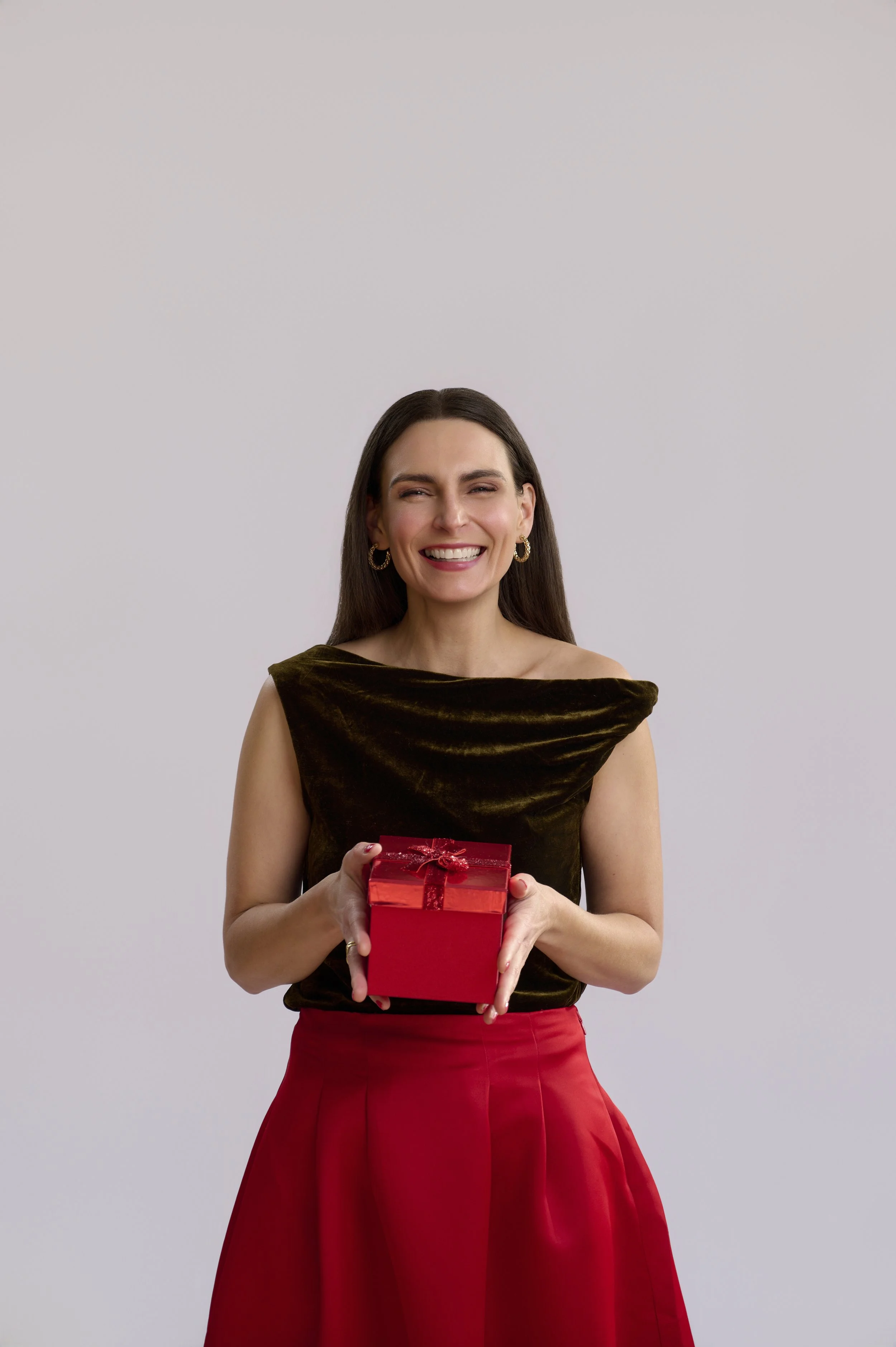 A woman smiling and holding a red gift box with a ribbon, wearing a brown sleeveless top and a red skirt, standing against a plain background.
