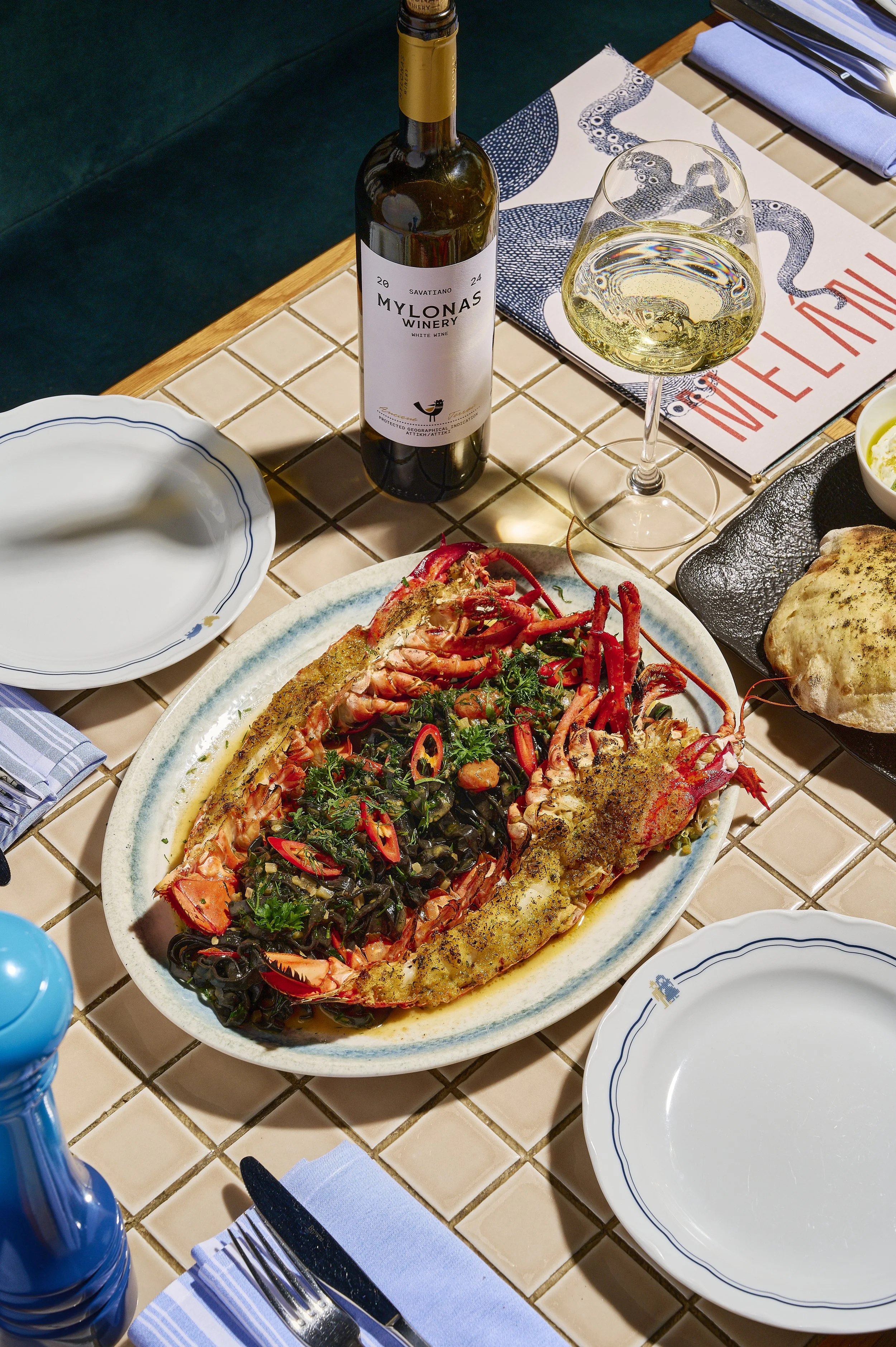 Lobster dish with herbs on a large oval plate, with a bottle of white wine, a glass of white wine, and place settings on a tiled table at a restaurant.