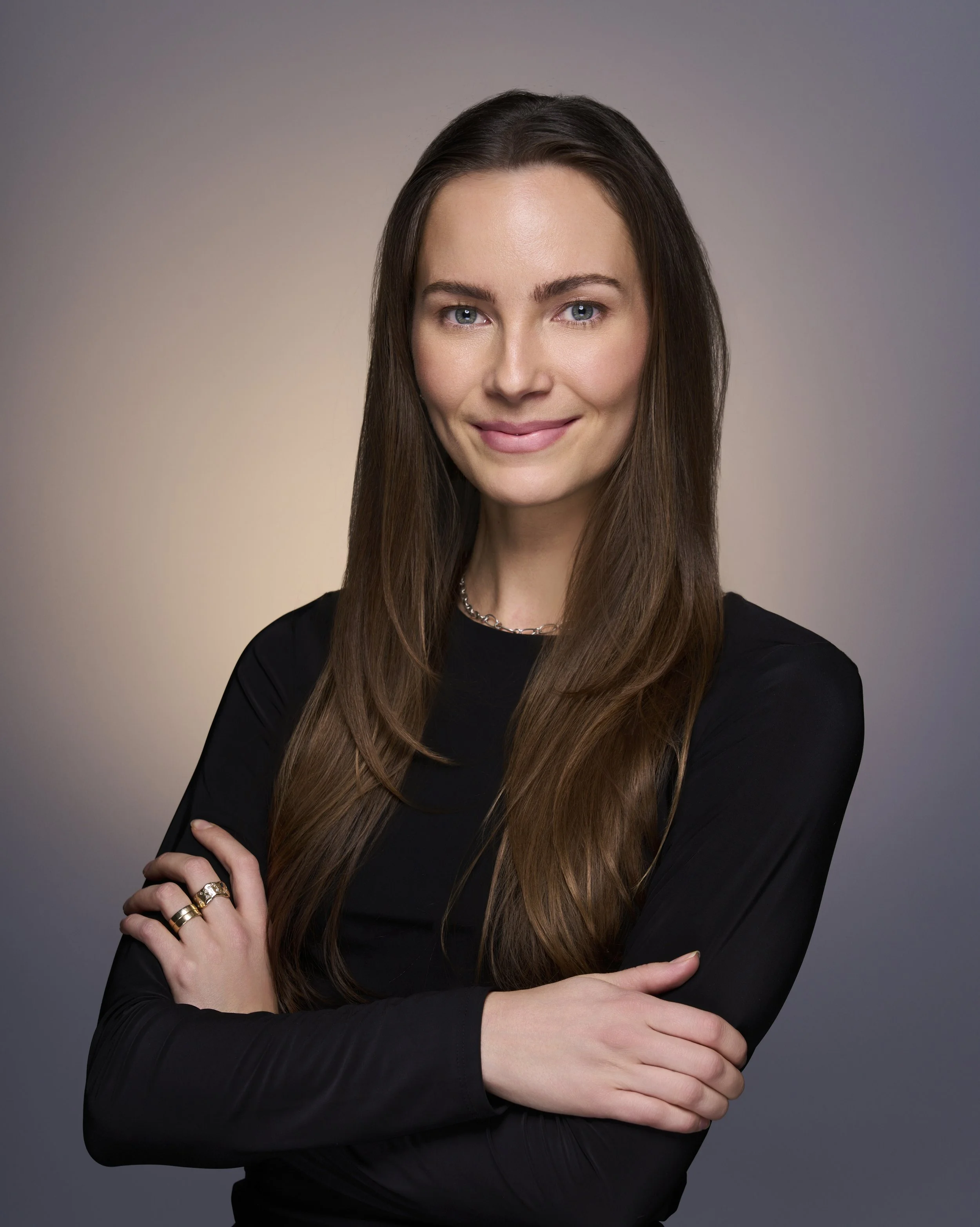 A woman with long brown hair wearing a black top, crossing her arms, smiling, against a gray gradient background.