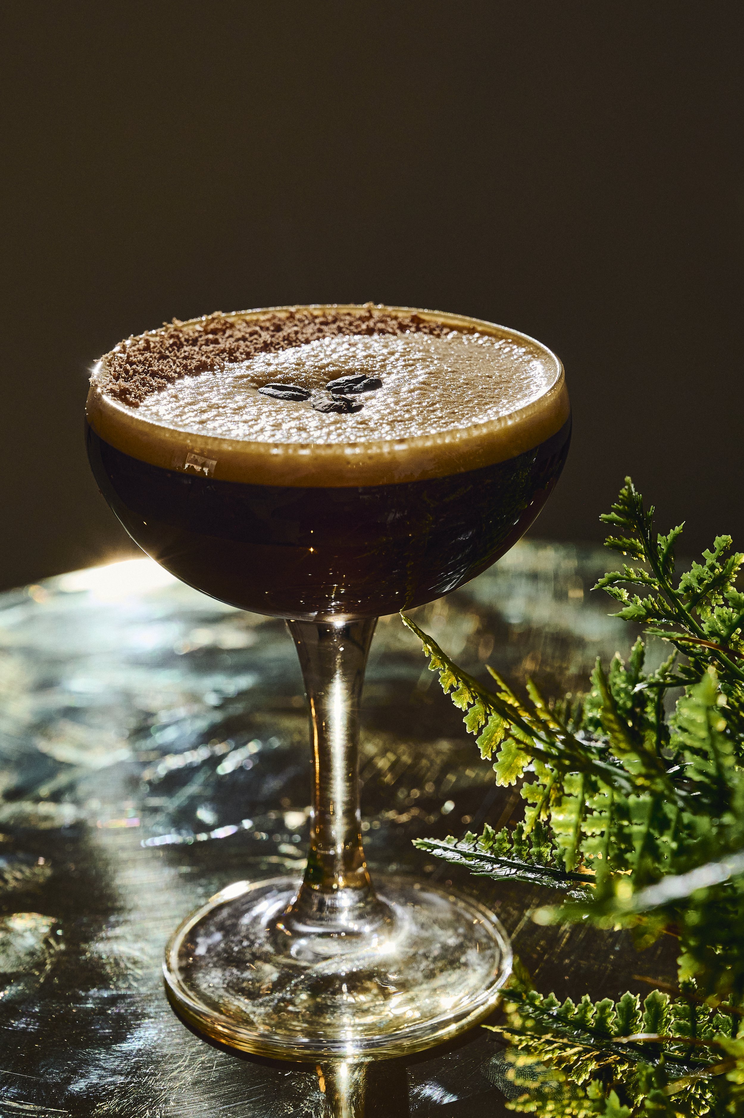 An espresso cocktail in a coupe glass with coffee beans on top, garnished with a chocolate rim, resting on a reflective surface with greenery beside it.