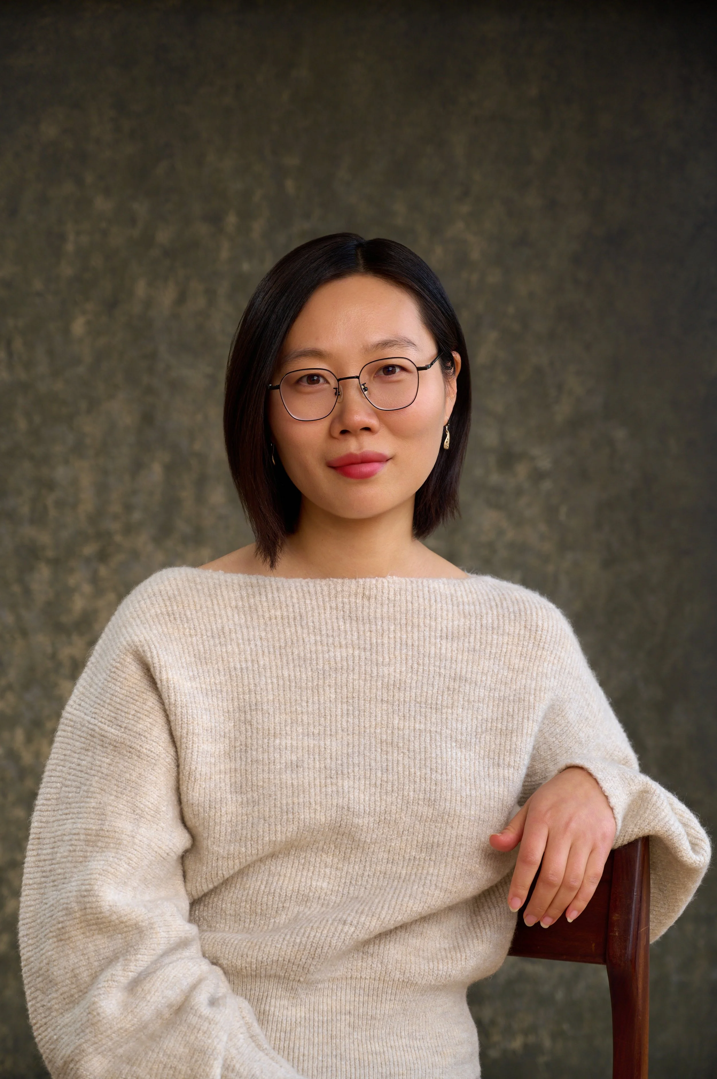 Portrait of a woman with shoulder-length dark hair, wearing glasses and a cream-colored sweater, sitting on a wooden chair, against a textured dark background.