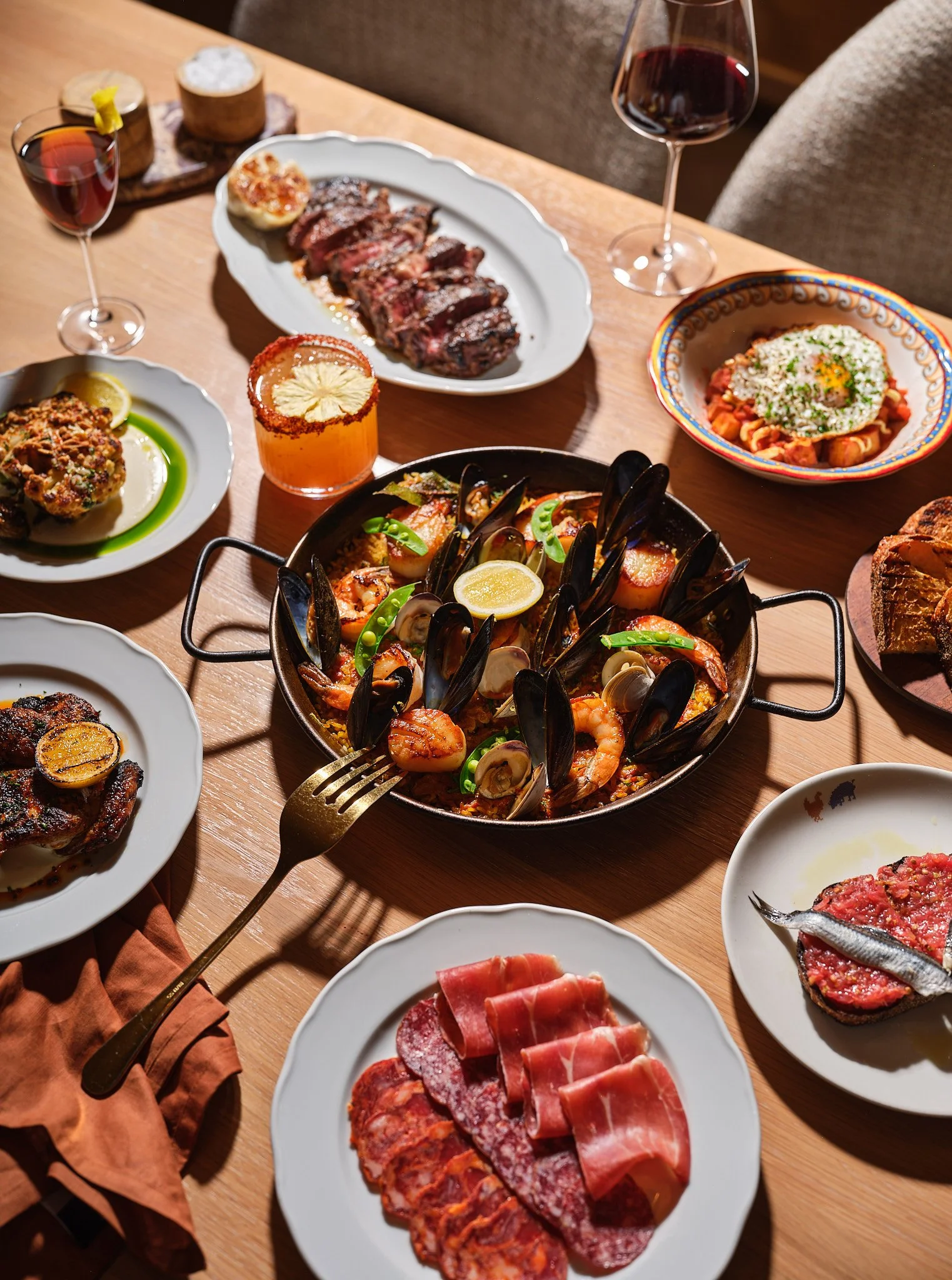 An Italian seafood and meat feast with dishes including a bowl of seafood with mussels, shrimp, and lemon, plates of cured meats, grilled meat, pasta, and a glass of red wine on a wooden table.