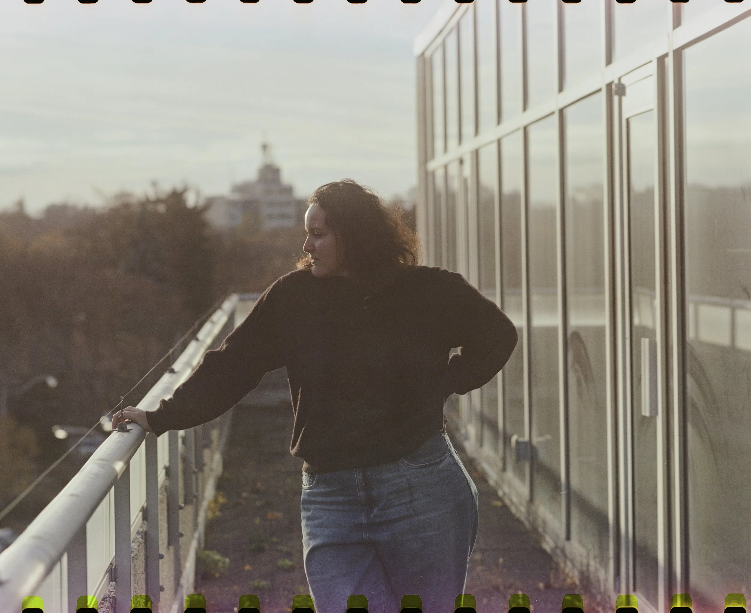A woman with curly hair standing on a balcony, leaning on the railing, with trees and buildings in the background during sunset.