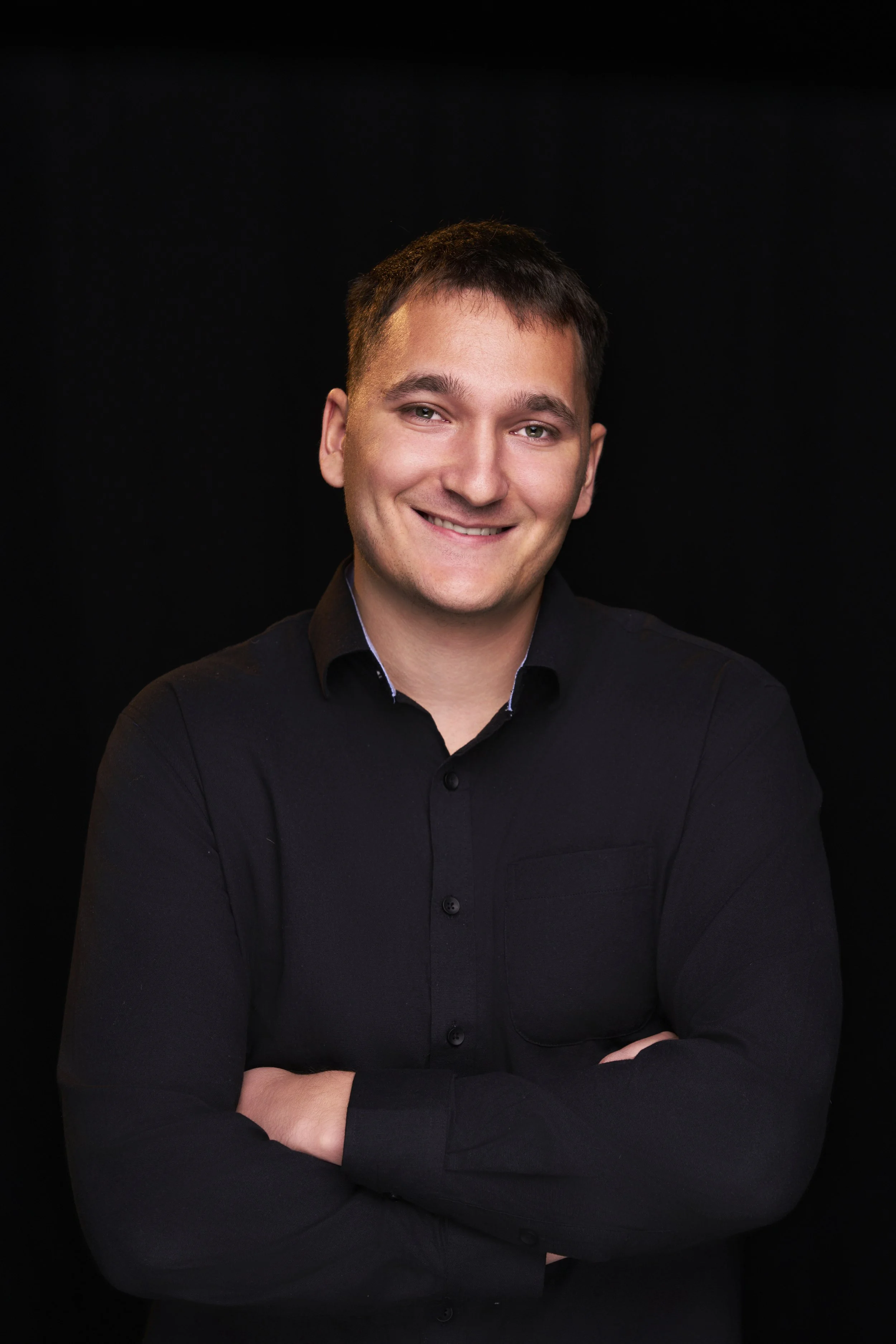 Portrait of a young man with short dark hair, smiling, wearing a black shirt, with arms crossed, against a black background.