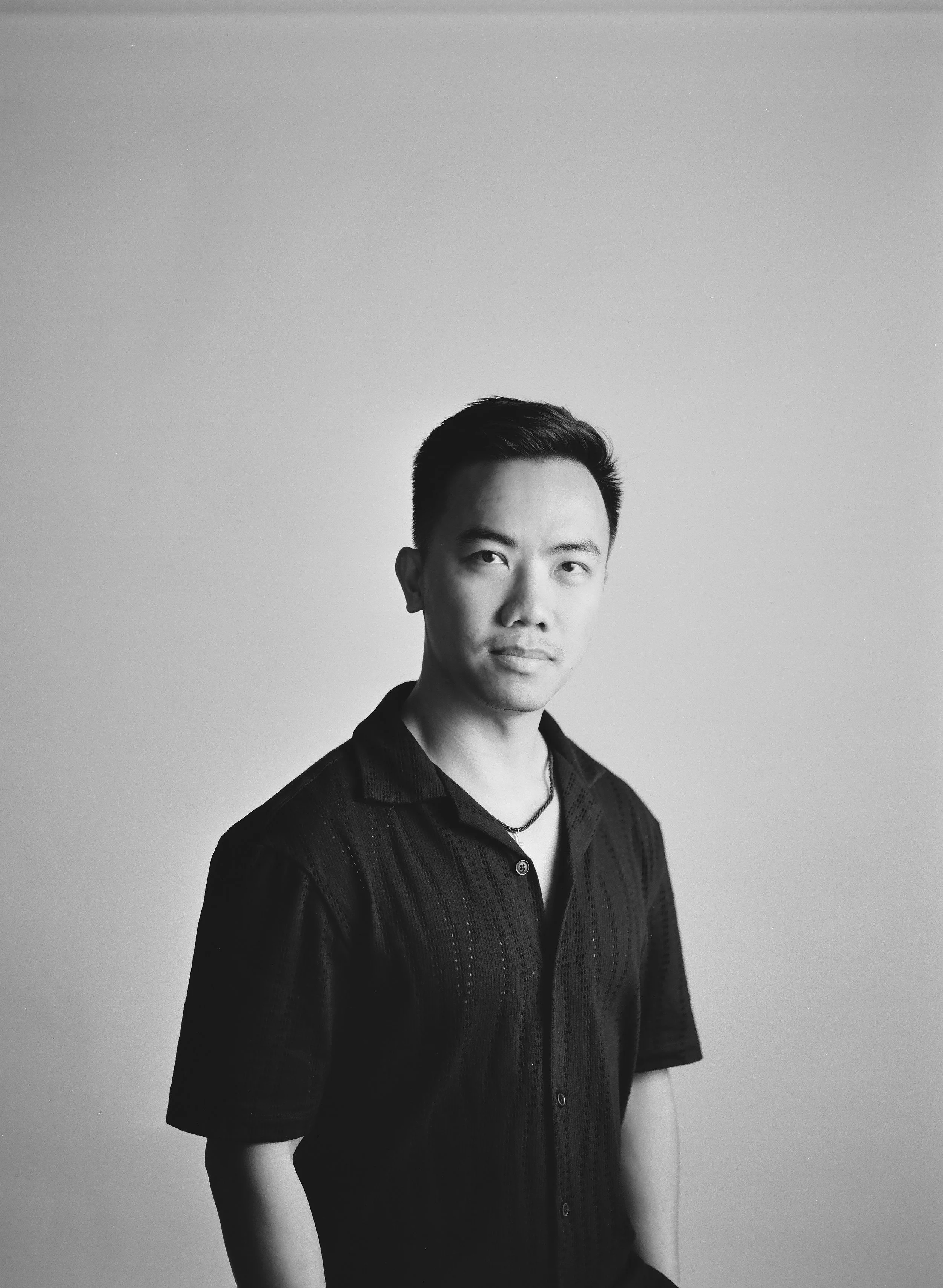 Black and white portrait of a young man with short dark hair, wearing a black shirt with a subtle pattern and a necklace, standing against a plain background.