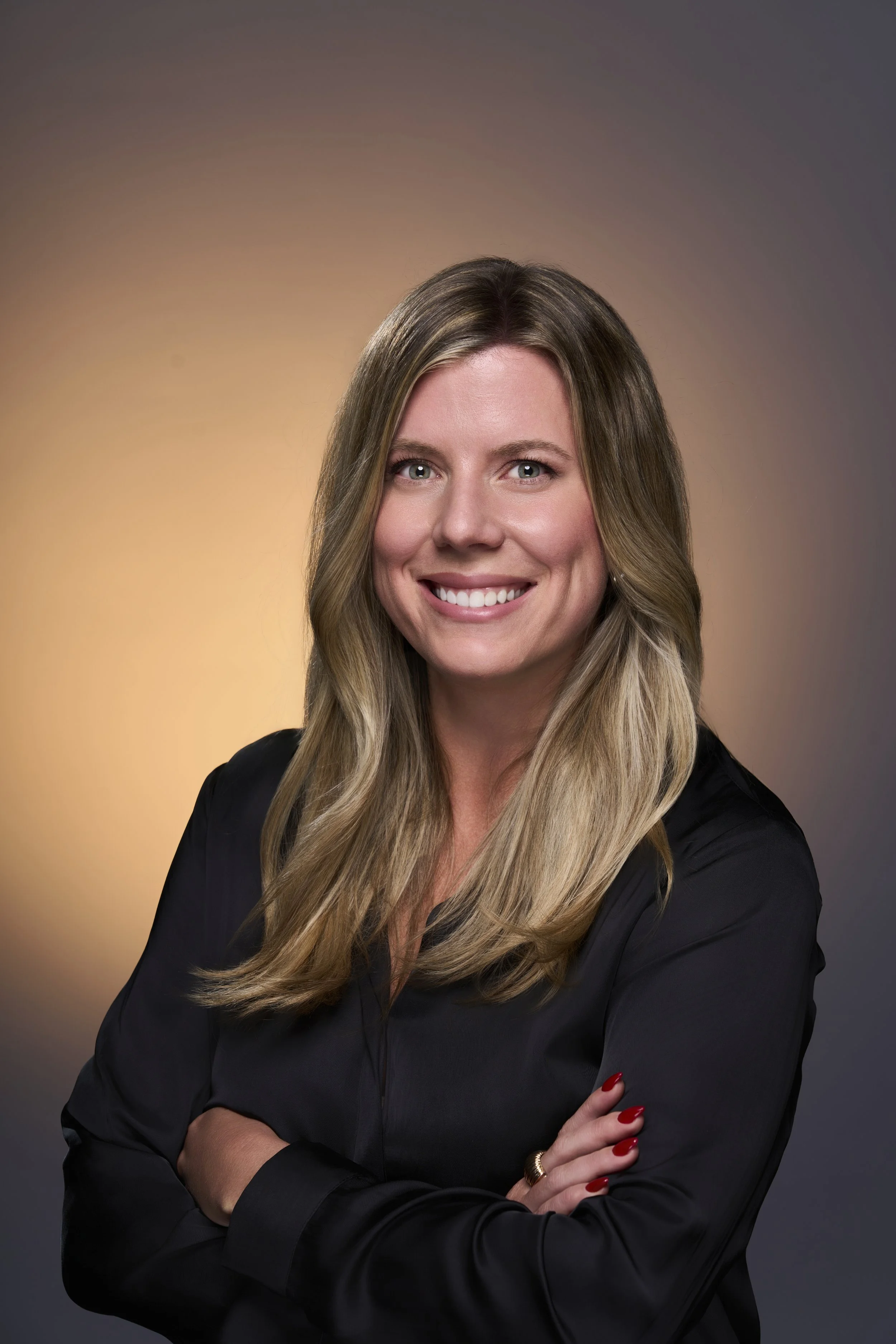 Professional headshot of a smiling woman with long blonde hair, wearing a black top, with crossed arms, against a gradient background.