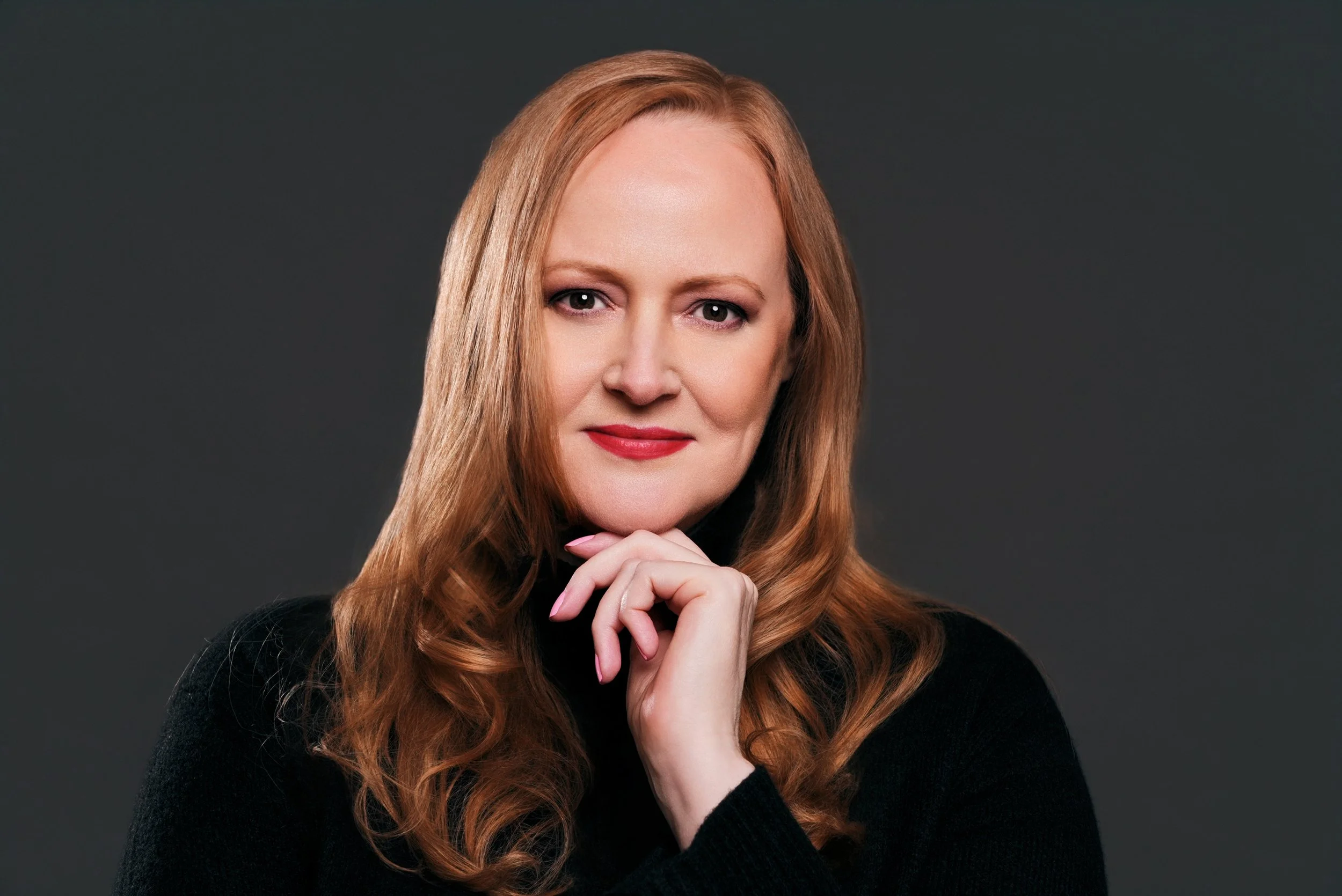 A woman with red hair, wearing a black top, posing with her hand under her chin against a dark grey background.