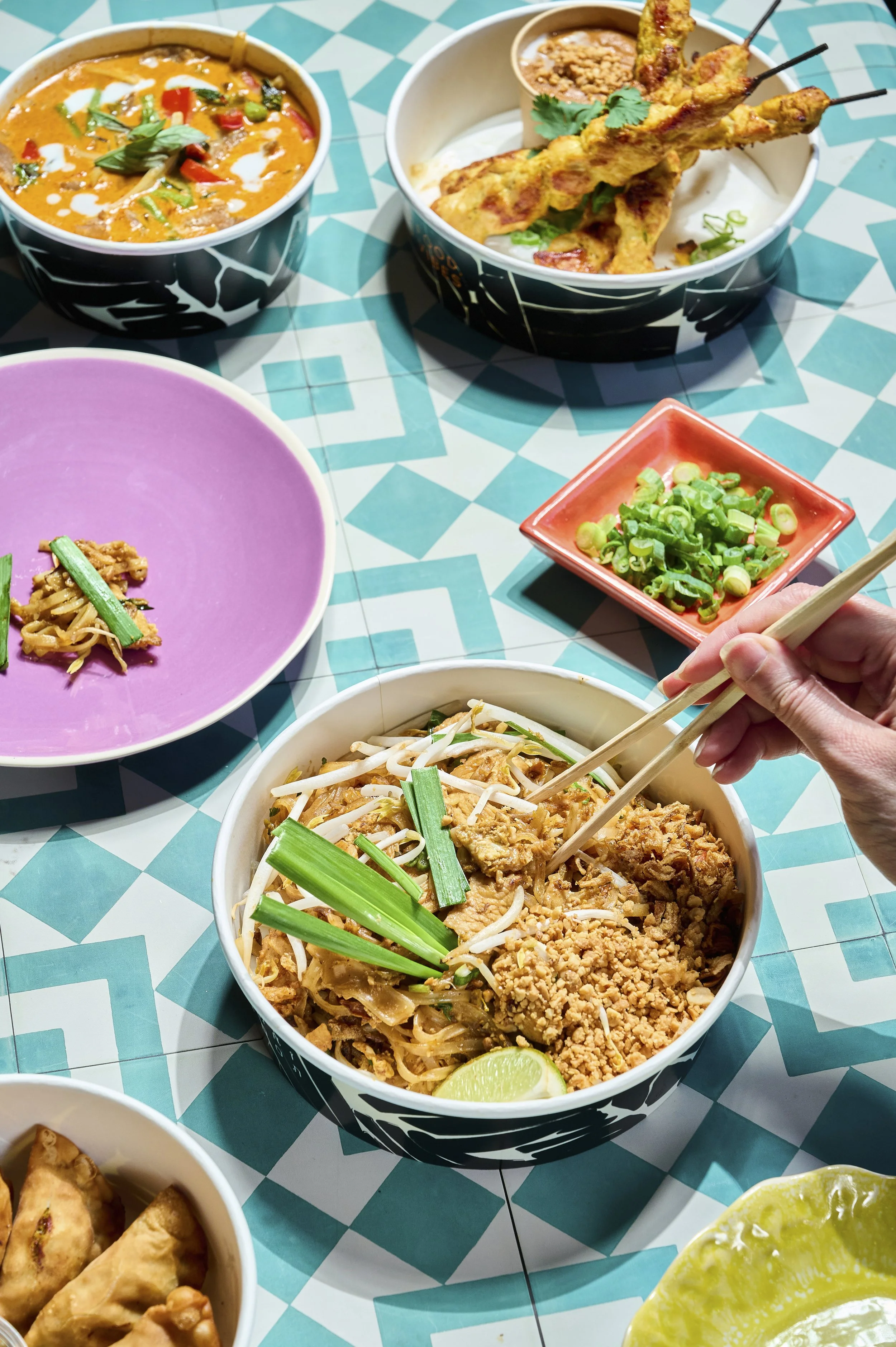 A variety of Asian dishes on a patterned table, including a bowl of pad thai with lime and green onions, a bowl of curry with garnishes, a bowl with skewers of grilled meat, and small bowls of chopped green onions and fried appetizers.