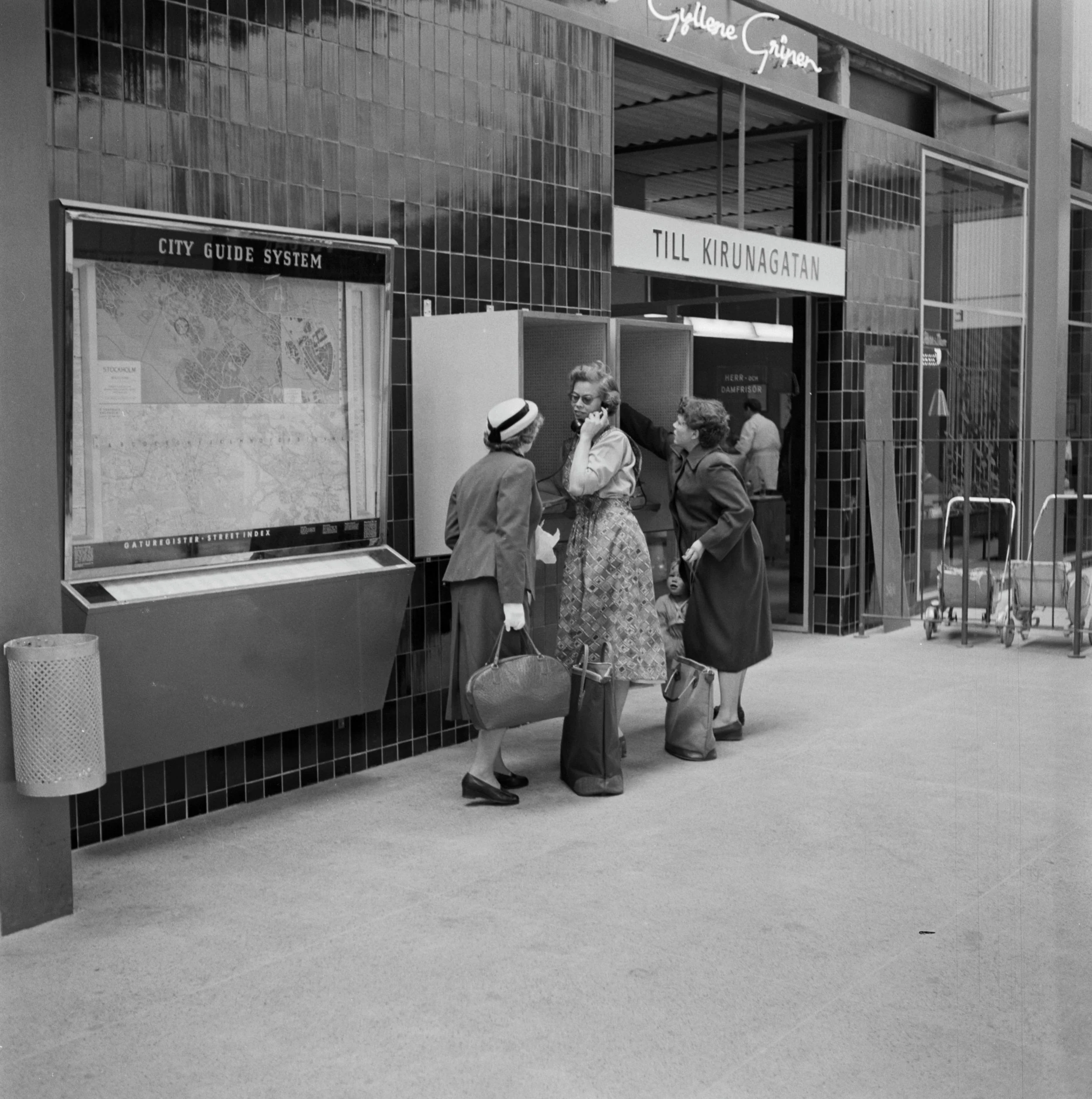 Tre kvinnor i biljetthallen på Vällingbys tunnelbanestation, vid telefonautomater. Bredvid dem syns en uppgång med texten "Till Kirunagatan" och en karta med rubriken "City guide system"