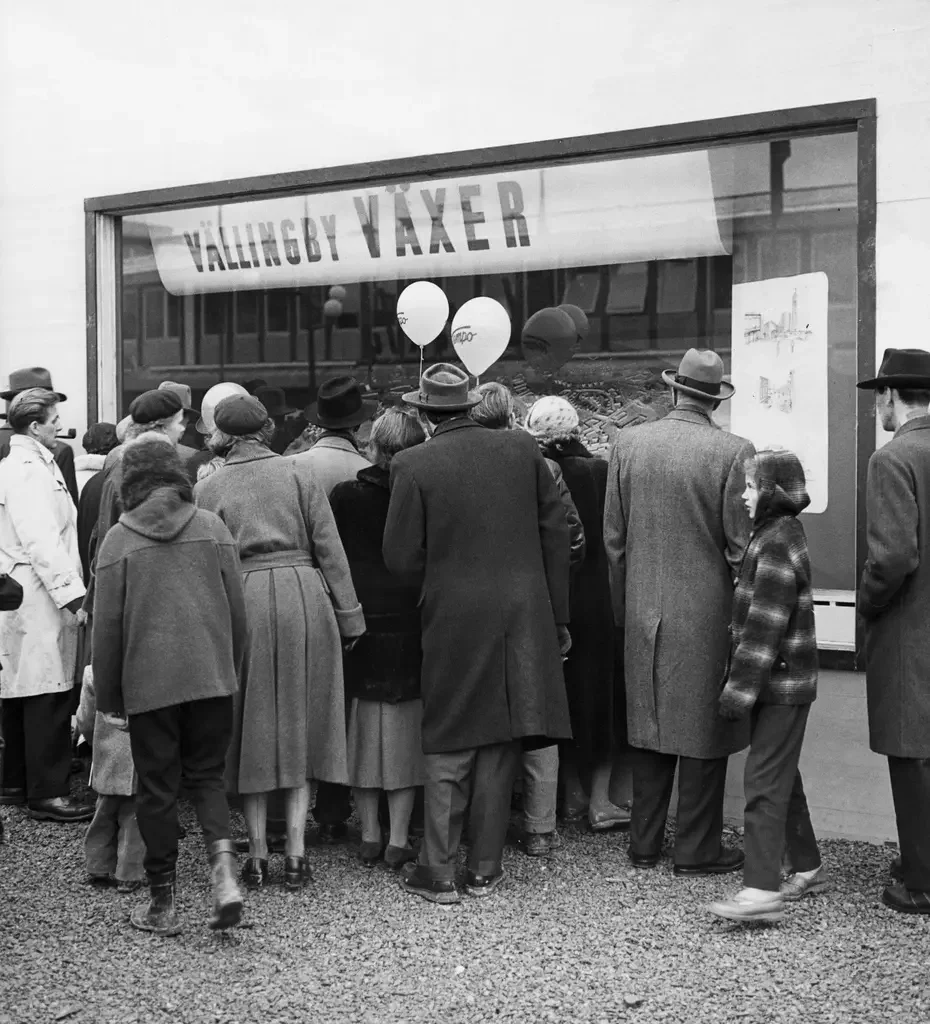 En grupp människor med rockar och hattar sedda bakifrån vid ett skyltfönster med banderollen "Vällingby växer"