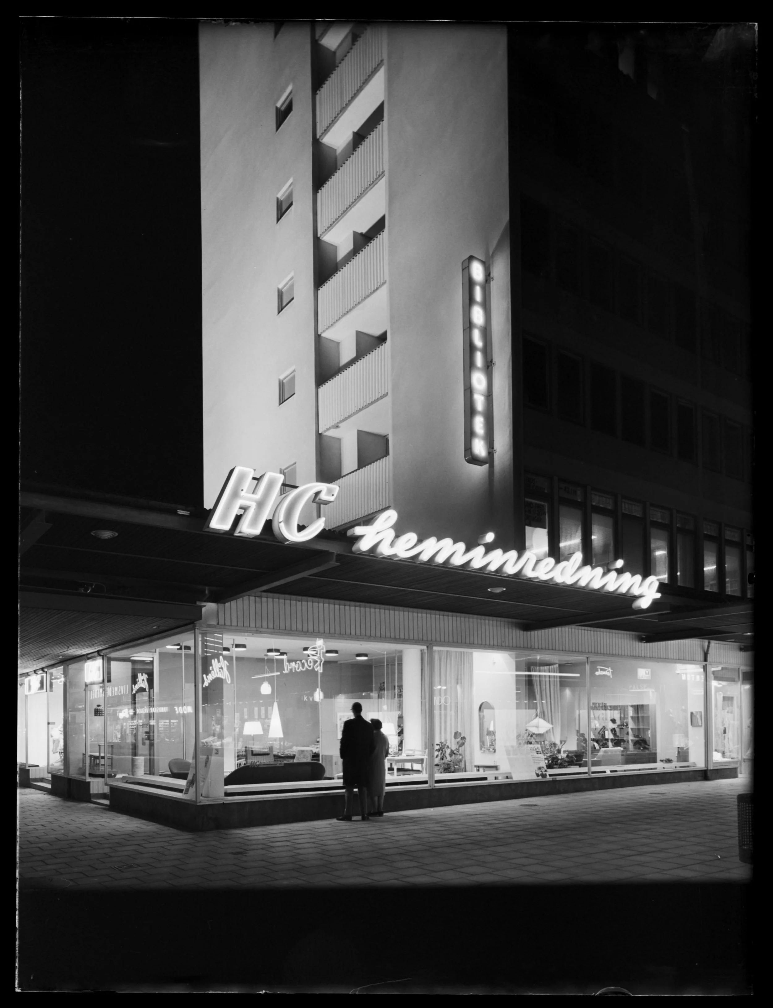 Två personer vid ett skyltfönster i centrum med neonskylten HC heminredning lysandes ovanför.