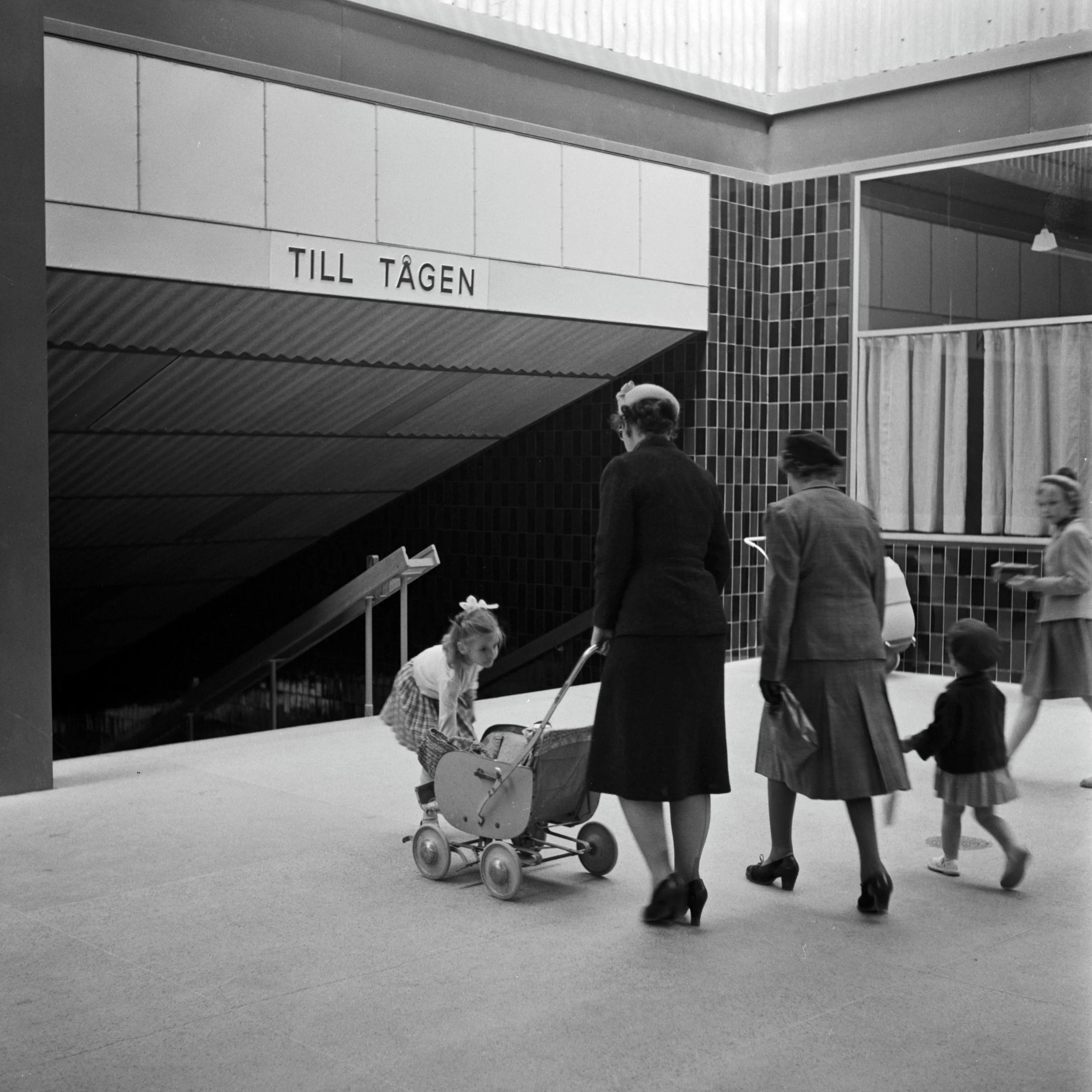 Två kvinnor med barn och barnvagn går mot tunnelbanenedgången, ovanför syns texten "Till tågen"