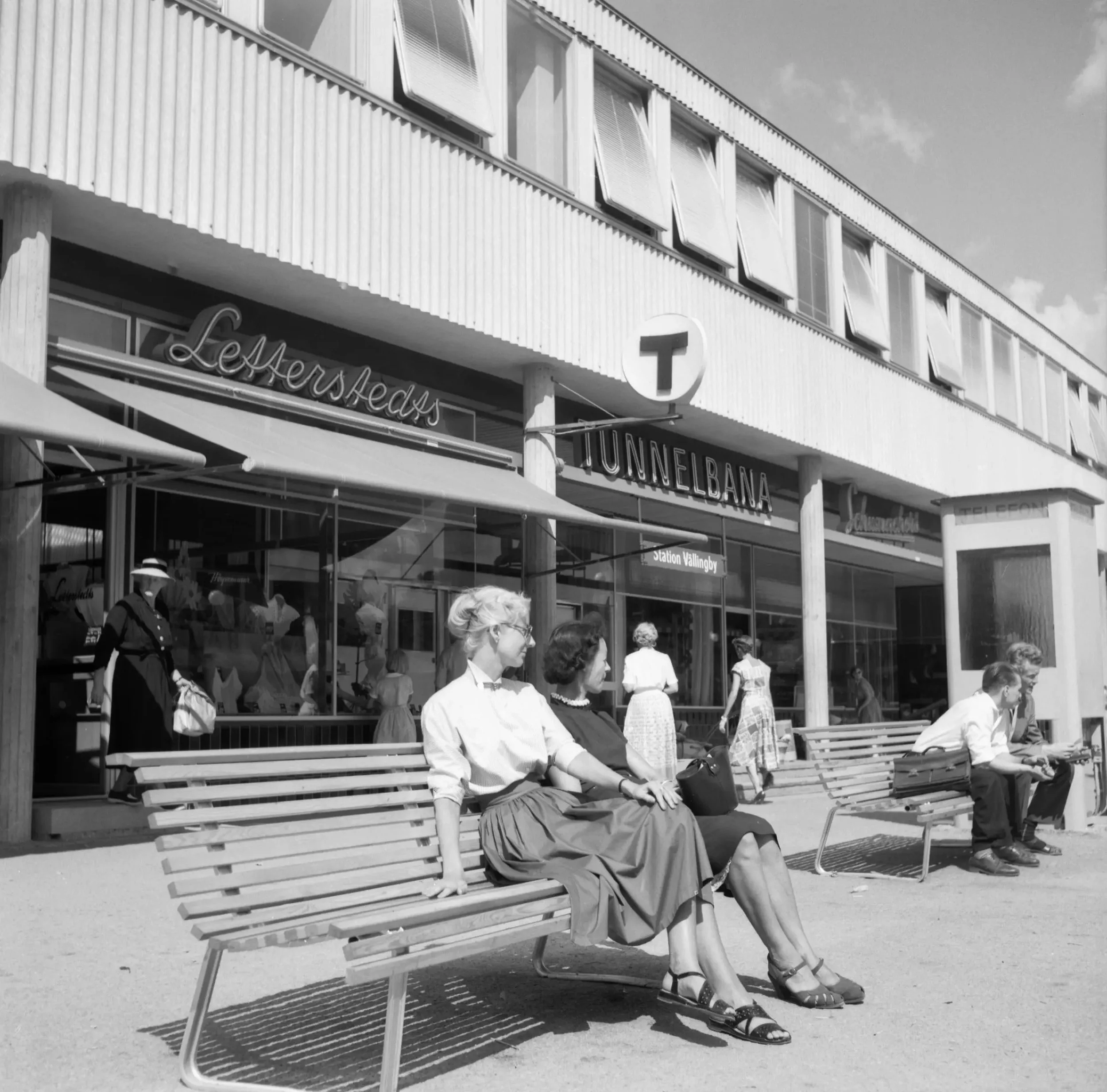 En bänk med två kvinnor och en bänk med två män i solen utanför Vällingbys tunnelbanestation.