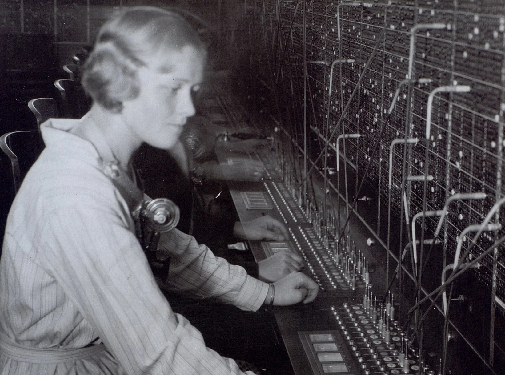 Kvinna vid telefonväxel, sedd från sidan med hörlurar runt halsen.