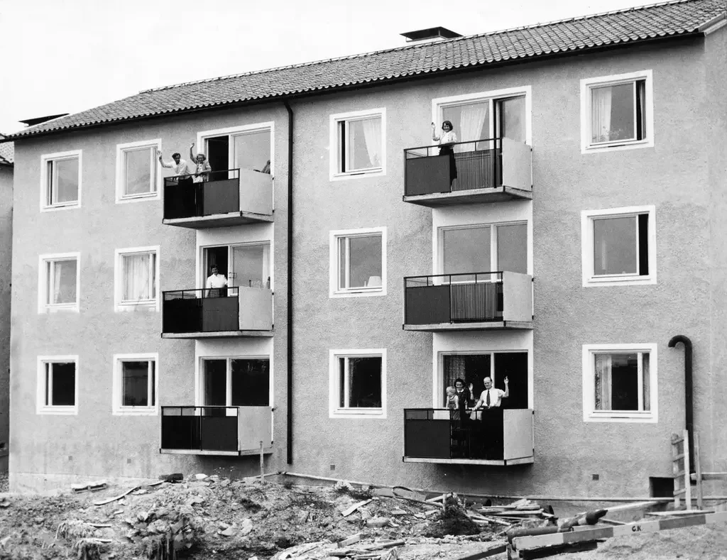 Svartvit bild av lägenhetshus i tre våningar med sex balkonger, på tre av balkongerna står personer och vinkar på sina lägenhetsbalkonger.